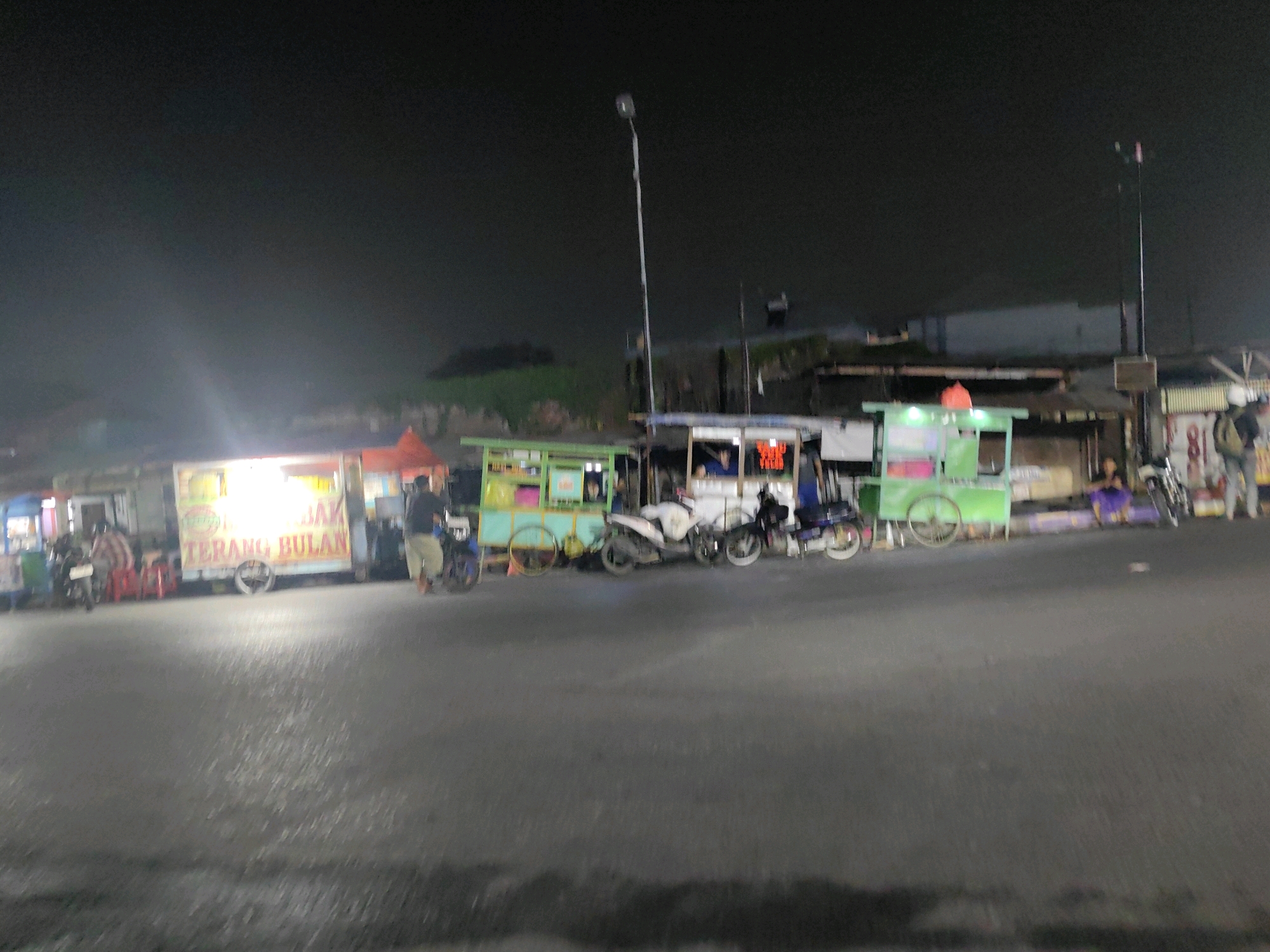 The image depicts a nighttime street scene in Keboansikep, Indonesia, featuring a row of food carts. In the foreground, the asphalt road is visible, with wet patches suggesting recent rain. Several food carts are parked along the side of the road. The cart on the far left has a white exterior with text that appears to be "BAK TERANG BULAN" in yellow and red lettering. To its right is a green food cart with a pink interior. Further down the row are other carts, one with a red awning. Parked near the carts are several motorcycles, including a white one, a dark-colored one, and a blue one. A street light stands tall on the left side of the frame, illuminating the area. People are visible in and around the carts, with one person standing near the "BAK TERANG BULAN" cart and others seated or standing near the green cart. The background shows the outlines of buildings and some vegetation under the dark night sky.