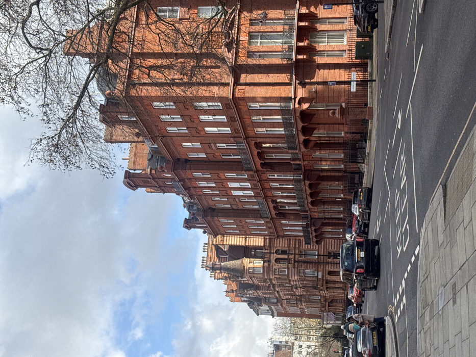 The image depicts a daytime street scene on Cadogan Square, London, England, under a partly cloudy sky. On the right, a row of ornate, multi-story red brick buildings with white-framed windows, intricate gables, and decorative balconies line the street. Bare tree branches extend into the upper right foreground. The asphalt road features a "KEEP CLEAR" box marking and dashed lane lines. A black cab (Hackney Carriage) is centrally positioned in the road, moving away from the viewer, while other vehicles (including a silver Porsche and several SUVs) are parked along both curbs and in traffic further down the street. On the left sidewalk, a single person in light-colored clothing stands near the parked Porsche, partially obscured. A street sign for "CADOGAN SQUARE" is visible on the right sidewalk.