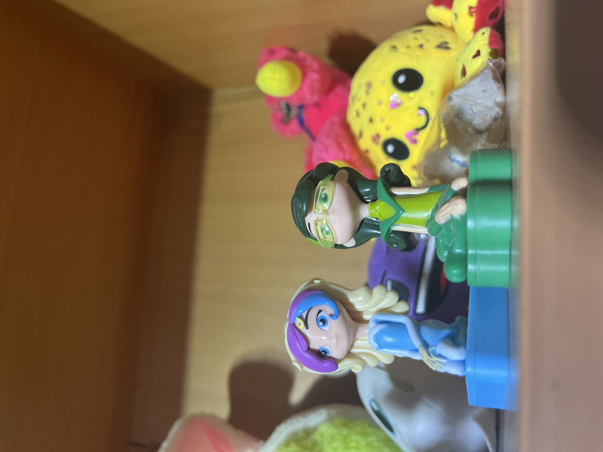 Two small figurines of cartoon characters are displayed on a wooden shelf. The figurine on the left has blonde curly hair with purple and blue streaks, blue eyes, and is wearing a blue dress. It is standing on a blue rectangular base. The figurine on the right has dark green hair, green eyes with large glasses, and is wearing a green and yellow outfit. It is standing on a green base. In the background, out of focus, are other toys including a plush yellow octopus with a smiling face, a pink plush toy with yellow accents, and a portion of a grey stuffed animal. The shelf is made of wood with a warm brown tone. The lighting suggests an indoor environment, possibly a room or display case. There are no people present in the image. The scene is static and does not depict any action. The location is not specified within the city of Ashkadar, Russia, as this is a generic indoor display.