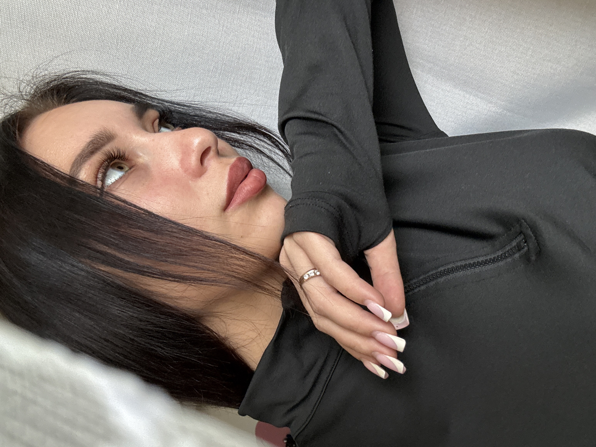 This media file captures a close-up, angled portrait of a woman lying down, looking upwards and to her right. She has long, dark hair, light-colored eyes, and well-groomed eyebrows. Her lips are full and appear to have a matte, reddish-brown lipstick. She is wearing a black long-sleeved top with a visible zipper detail on the chest area. Her right hand is prominently featured near her face, adorned with a ring featuring three small clear stones. Her nails are long, manicured with a white French tip and an additional diagonal white design on a light pink base. The background consists of a plain, light grey, textured fabric, suggesting she is lying on a bed or couch. The lighting is soft and diffused, indicating an indoor setting. The woman's gaze suggests a pensive or relaxed mood. There is no visible text in the image. The scene is set in Chaikovsky, Russia.