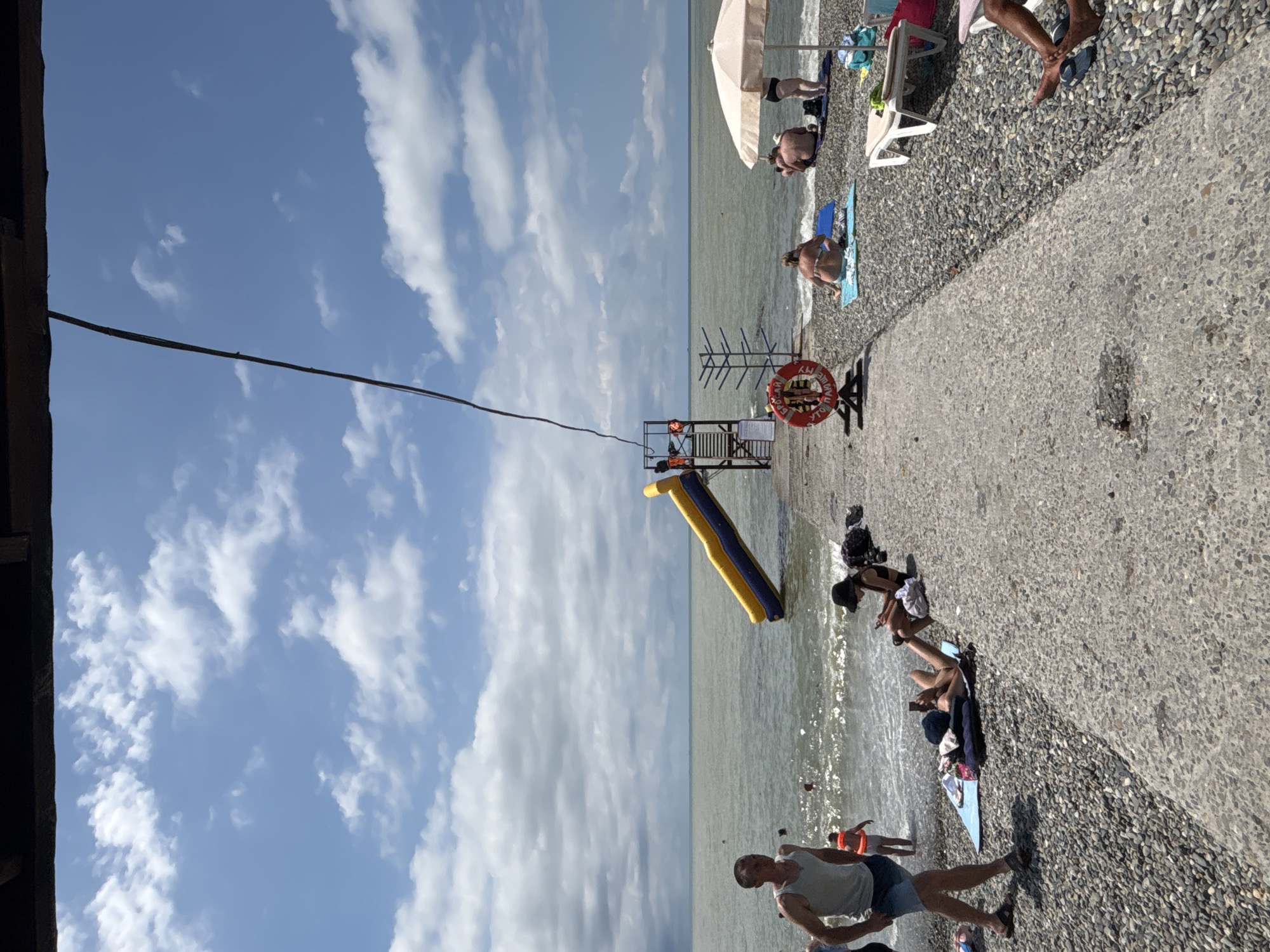 This image depicts a sunny day at a beach in Sochi, Russia, likely a popular tourist area due to the presence of an inflatable water slide extending from a concrete pier into the sea. The pier itself is made of concrete and gravel, with numerous people scattered across it, sunbathing, or relaxing.
On the pier, a vibrant yellow and blue inflatable slide is a prominent feature, positioned to allow access into the water. Adjacent to the slide is a lifeguard stand with a red and white life preserver in the foreground, suggesting safety measures are in place. The sea is calm with gentle waves lapping at the shore.
The beach is covered in grey pebbles, with several beach towels and umbrellas laid out. People are seen in various states of relaxation: some are sunbathing on towels, others are sitting or lying down. A few individuals are in the shallow water, while others are walking along the shore.
In the foreground, a man in a light-colored tank top and dark shorts is walking, while further down the pier, a woman in a black swimsuit and hat is using her phone. Another person is lying on a towel, also engaged with a mobile device. In the background, more beachgoers are visible under umbrellas and on towels. The sky is a clear blue with scattered white clouds, indicating pleasant weather conditions. The overall scene conveys a relaxed and leisurely atmosphere typical of a summer beach day.