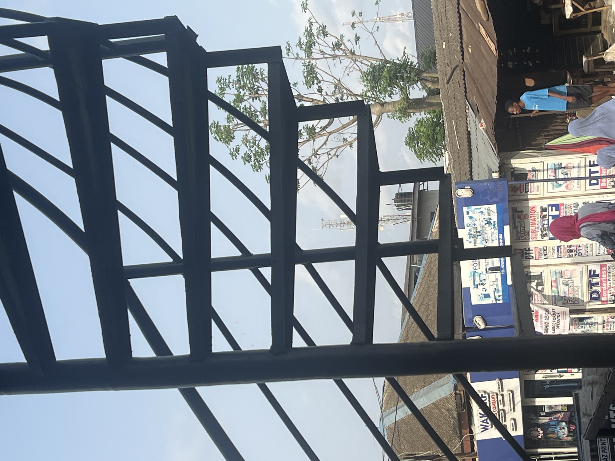 The image shows a street scene in Lagos, Nigeria, viewed through a metal structure (possibly a staircase or railing). In the background, there are buildings and shops with various signage, including advertisements for printing services (DTF, Sublimation, Large Format Printing), one with a job posting for a "Graphics Designer/Large Format Operator". People are visible in the background, including one wearing a blue shirt. Vegetation is seen through the metal framework.