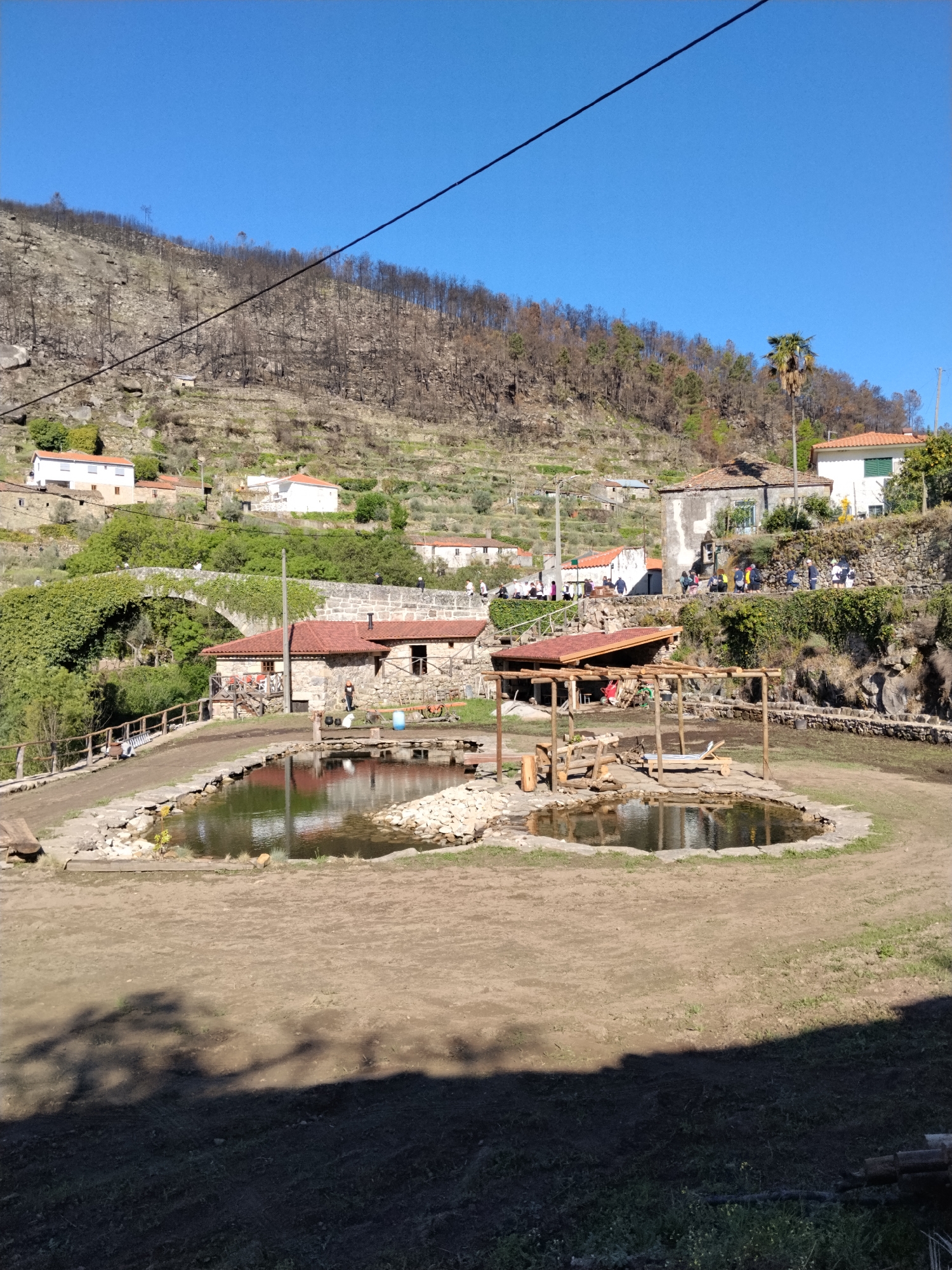 The image shows a rural scene in Tarouca, Portugal. In the foreground, there are two artificial ponds or pools enclosed by a dirt area. Nearby, there's an open wooden structure, possibly a pergola or shelter.
In the mid-ground, there are stone buildings with red tile roofs, a stone bridge covered with greenery, and a stone wall. Several people are visible on a path, near the stone buildings.
In the background, a hillside covered with vegetation and some buildings is visible under a clear blue sky. The vegetation appears to be partially burned. A utility line stretches across the upper part of the image.