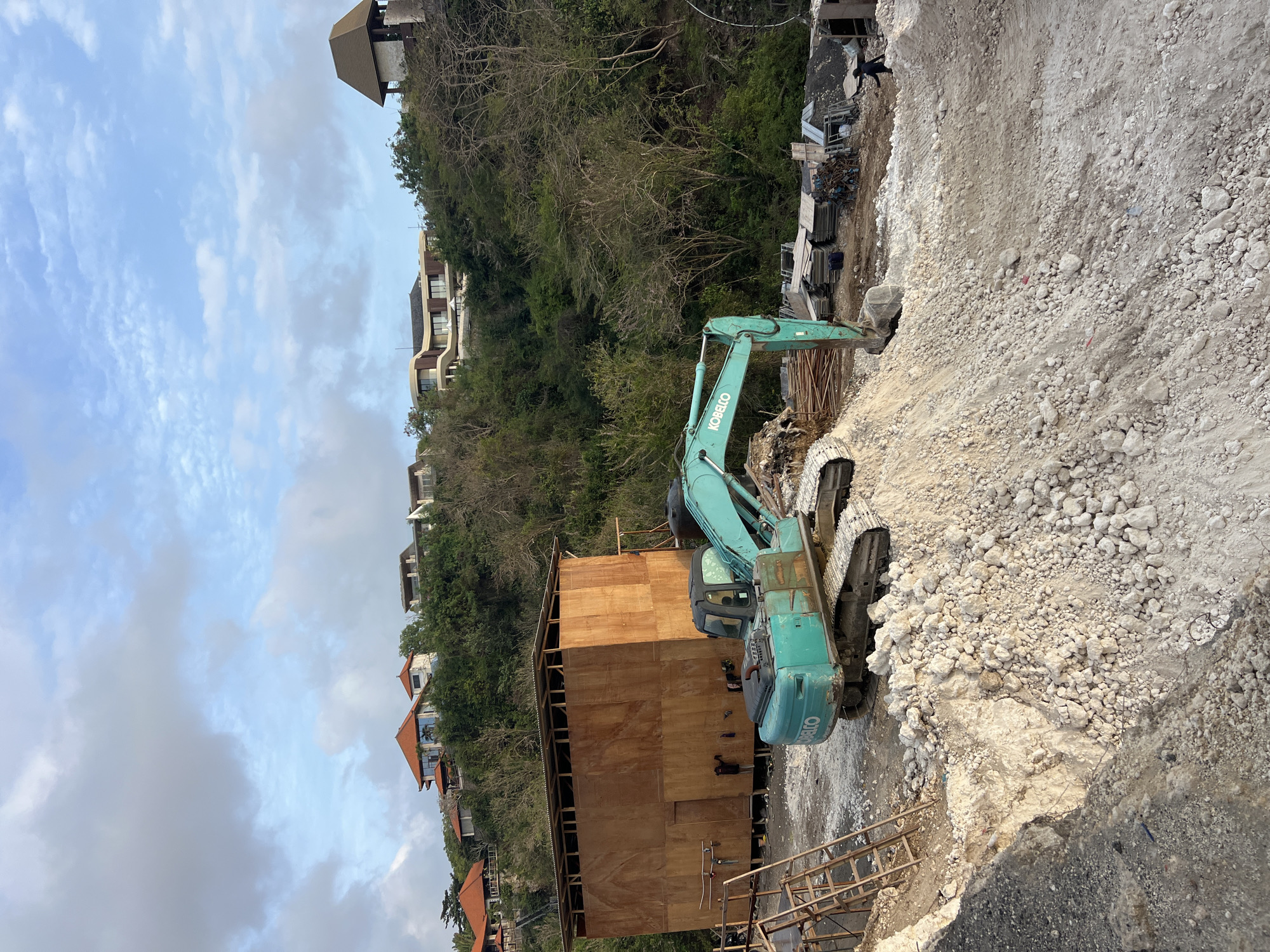 In Kuta Selatan, Indonesia, a construction site is depicted on a lush, green hillside. The foreground is dominated by a large pile of light-colored rubble and dirt, with a portion of a construction excavator, painted teal and branded "KOBELCO," partially submerged in the debris. To the left, a wooden structure with a corrugated metal roof is under construction, with scaffolding and construction materials nearby. Behind this, the hillside rises steeply, covered in dense vegetation and trees. Higher up the hill, several modern houses with red-tiled roofs and balconies are visible, nestled amongst the greenery. The sky above is a light blue, with scattered white clouds. The overall scene suggests ongoing development in a tropical coastal area.