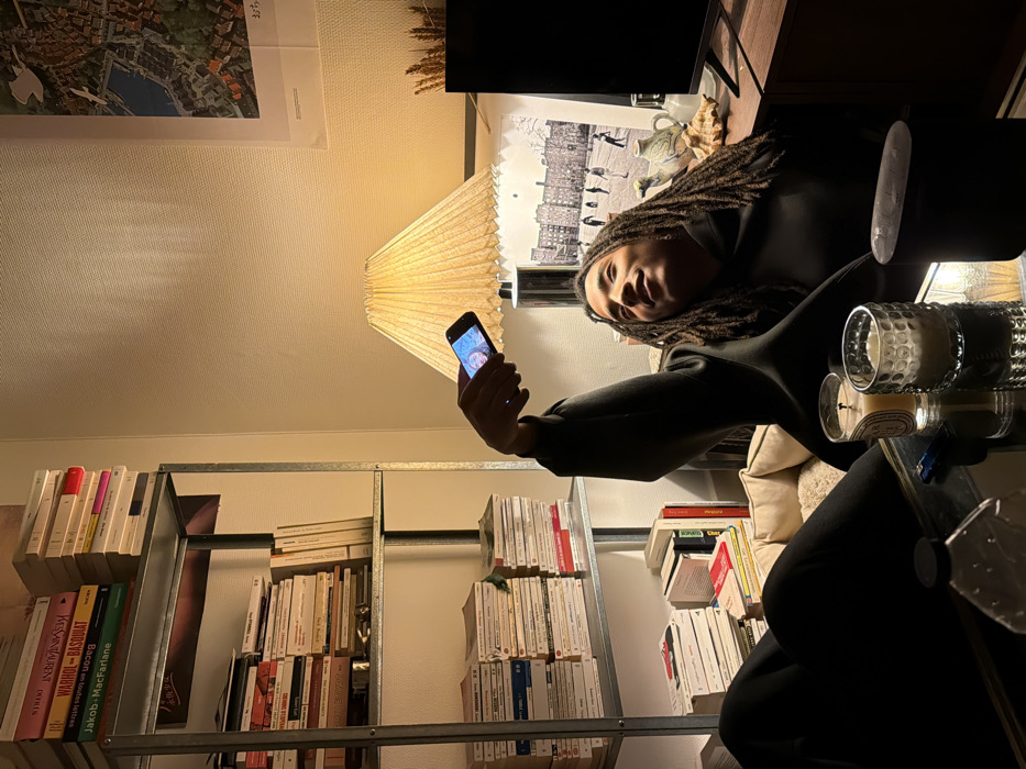 A person with dreadlocks holds up a phone, taking a selfie. They are seated next to a glass-topped table with candles, near a tall bookshelf filled with books. A pleated lampshade hangs overhead, casting a warm glow. Behind the person, there is a framed black-and-white picture and other objects on a shelf. A framed landscape picture is visible on the wall above the bookcase.