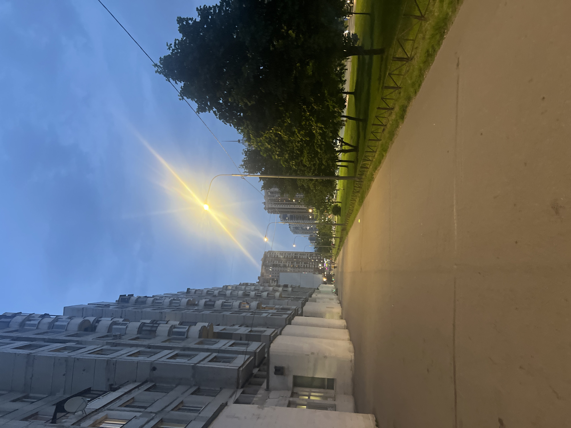This image depicts an urban street scene during twilight or early night in Saint Petersburg, Russia.
On the left, a long, multi-story residential building, consistent with Soviet-era panel architecture, extends into the background. The building is grey, featuring numerous windows and recessed balconies, with at least one visible satellite dish on an upper floor. The lower section of the building is white with distinct, vertically elongated, slightly angled supports or columns.
A wide, dark grey paved walkway or minor road occupies the central and foreground area, extending into the distance. It appears mostly clear, with some faint marks or stains.
To the right of the walkway, a narrow strip of green grass is bordered by a low, dark, segmented metal fence. A line of deciduous trees with dense, dark green foliage stands within this grassy area. Beyond the trees, the faint outlines of vehicles with illuminated lights suggest a parallel road or street.
The scene is illuminated by several streetlights emitting a bright yellow-orange light, with a prominent light in the mid-left foreground producing a distinct lens flare. The sky above is a deep blue, indicative of dusk or early night, with visible cloud cover.
In the mid-distance on the walkway, a few indistinct human figures are visible. Further in the background, additional multi-story residential buildings are apparent, some with illuminated windows. Crucially, a tall, slender, architectural spire is visible in the distant background, rising above the other buildings and trees. This spire is identifiable as the **Lakhta Center**, a landmark skyscraper in Saint Petersburg, confirming the location.