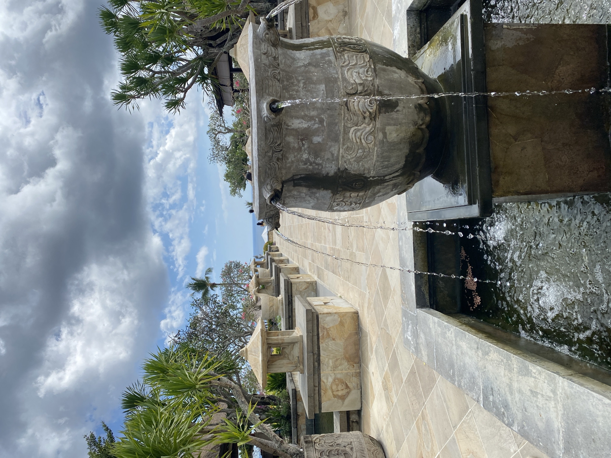 An outdoor, tropical scene in Kuta Selatan, Indonesia, features a water fountain as the central subject. The fountain is a large, ornate stone vessel with four spouts from which water cascades into a stone basin. The basin is filled with dark water, and some brown leaves or debris are visible floating on the surface. The fountain is placed on a tiled patio, with a pathway of light-colored square tiles extending into the background.
Along the pathway, a series of similar stone structures are arranged in a line, each topped with a decorative urn or a small lantern-like architectural element. These structures are made of large, irregularly shaped stone blocks. Lush green foliage, including palm-like trees and flowering bushes, lines the pathway on either side.
In the background, the ocean is visible under a sky filled with dramatic, dark grey clouds interspersed with patches of blue sky and lighter white clouds. A few distant figures can be seen near the edge of the property, suggesting a resort or hotel setting. The overall impression is one of serene, tropical landscaping with flowing water elements.