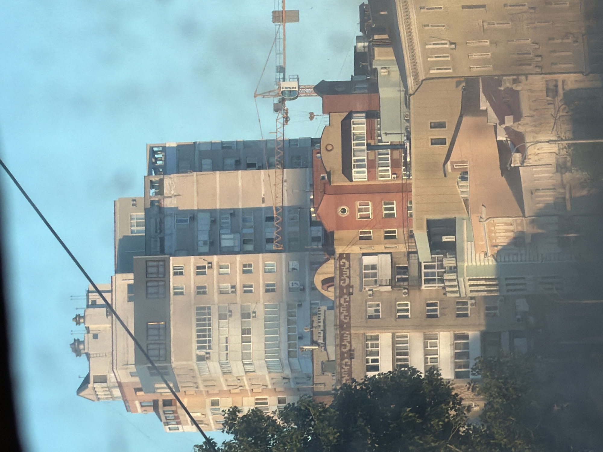 The image shows a cluster of multi-story buildings under a clear blue sky, with a construction crane visible in the mid-ground. The buildings vary in architectural style and color, ranging from light beige and grey to salmon pink. Some buildings feature prominent balconies and numerous windows. A large tree partially obscures the left side of the scene. The lower portion of the image is blurred, suggesting it may be a reflection or a result of motion. The scene is consistent with an urban environment, and the style of architecture is typical of residential buildings found in Rostov-on-Don, Russia.