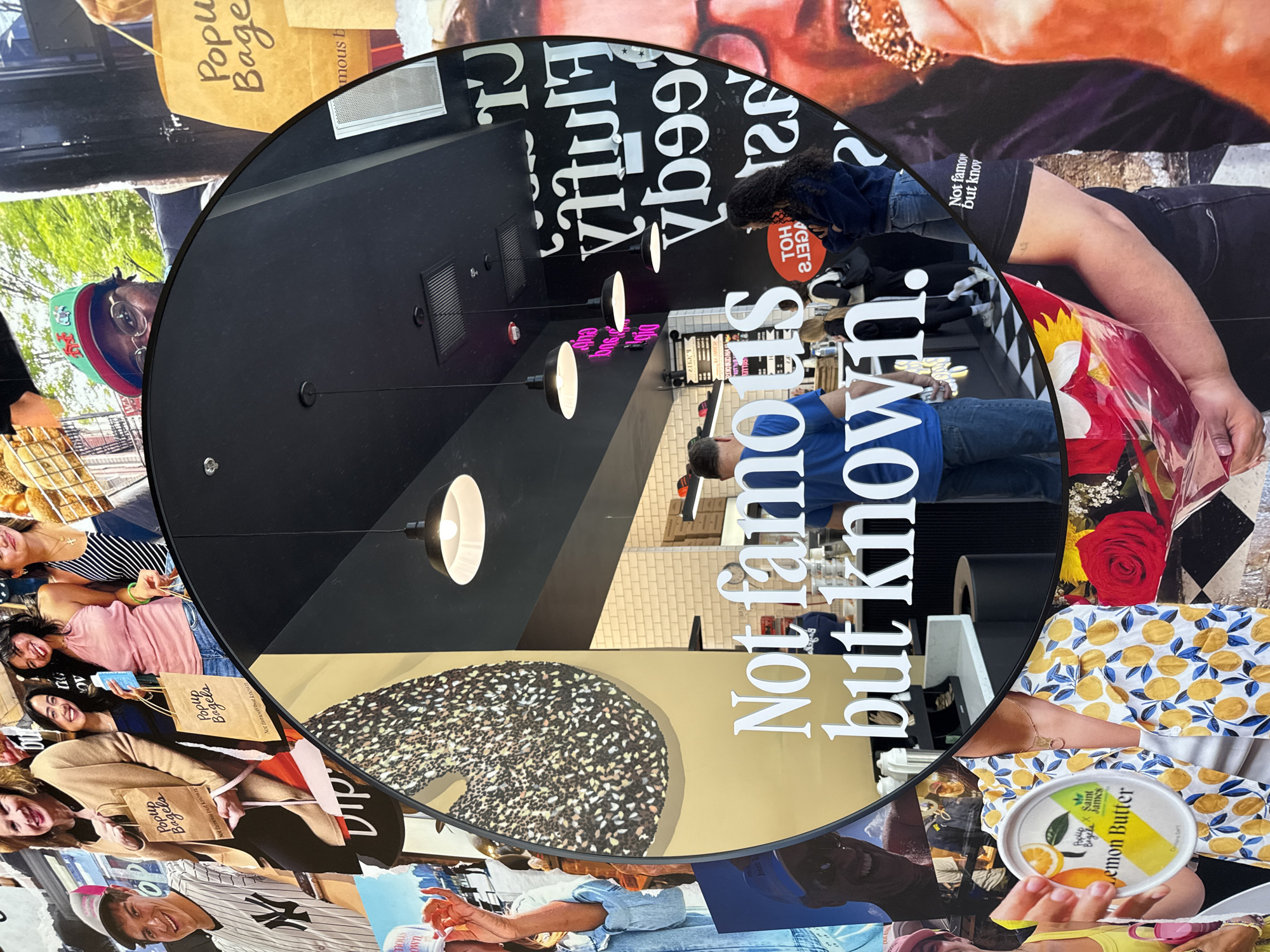 The image shows a circular display with a reflective surface, reflecting the interior of a business. The background of the display is a collage of various images including people holding "Popup Bagels" branded bags, a person wearing a New York Yankees baseball cap, and a jar of "Saint James Lemon Butter." The reflection inside the display shows a well-lit interior with pendant lights, signage with the reversed text "CT Hutta", and a phrase "Not famous but known" in reverse. People appear to be inside the business. The ceiling is black, and walls are tiled in white.