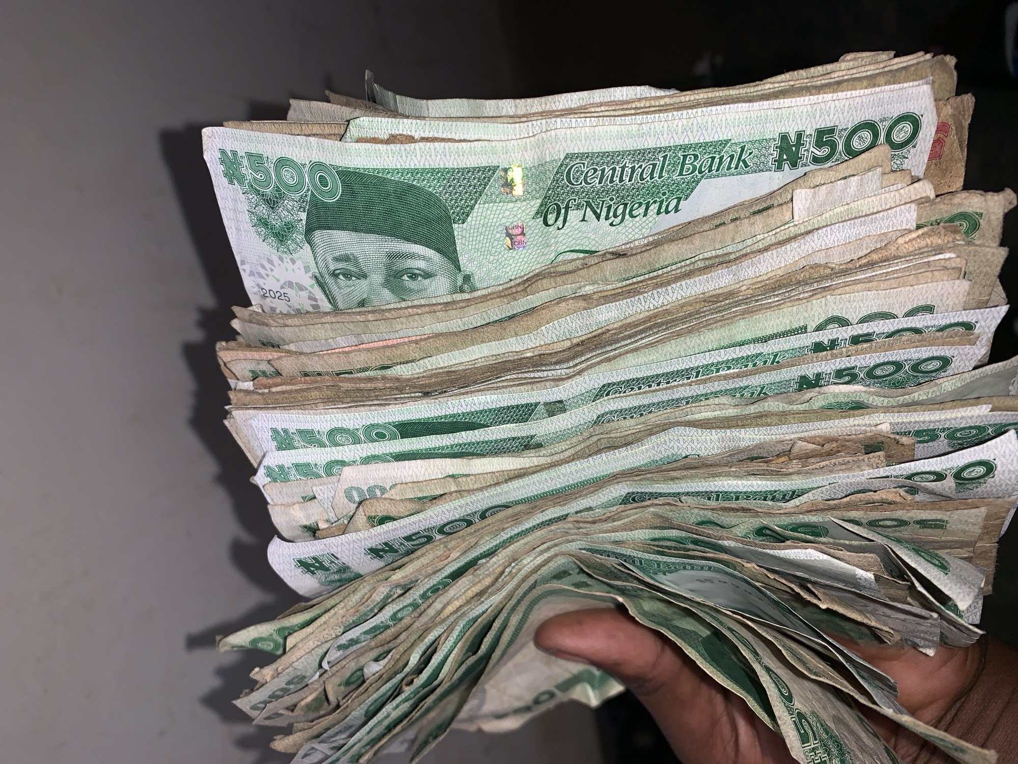 A close-up shot reveals a stack of Nigerian 500 Naira notes held in a person's hand against a muted grey background. The notes are primarily green and white, with the "Central Bank of Nigeria" prominently displayed. A portrait of a man wearing a traditional hat is visible on the notes, and the denomination "500" is printed on each. The notes appear somewhat worn and crumpled. The person's hand is dark-skinned and holds the stack of currency firmly. The environment is indistinct, offering little additional information. The location is not discernable from the image.