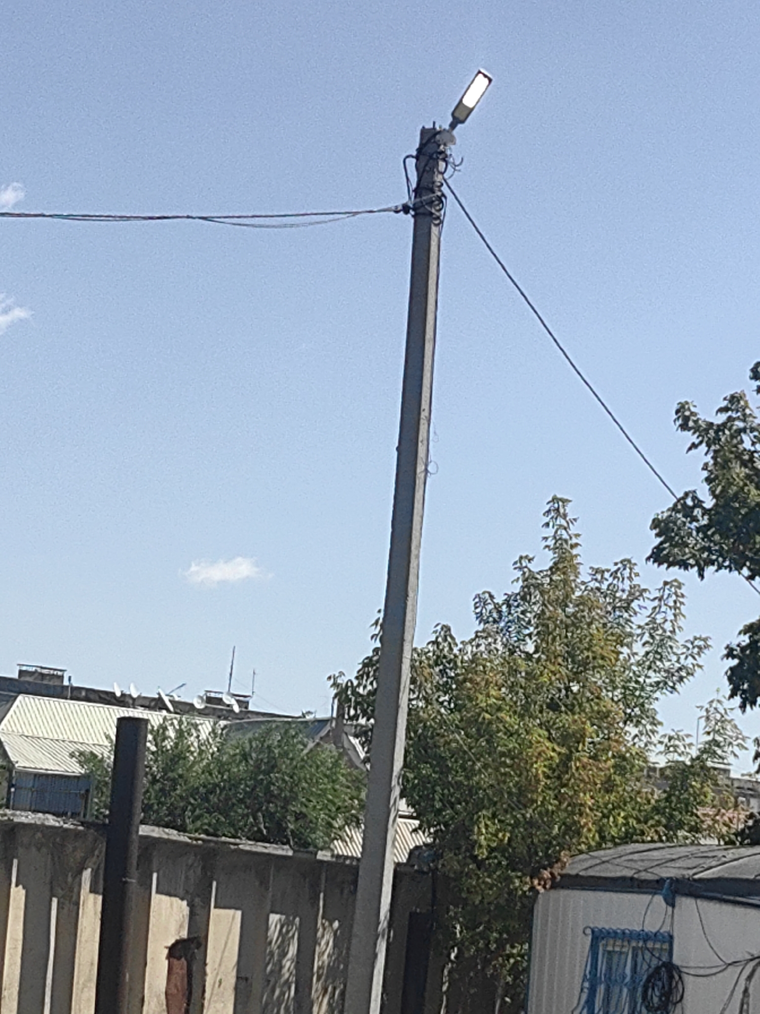 The image shows a street lamp on a concrete pole against a clear blue sky in Shymkent, Kazakhstan. The lamp is a modern, elongated LED fixture. Wires are strung from the pole, disappearing into the upper left and right corners of the frame. Below the sky, a wall made of concrete blocks is visible, with vertical shadows cast upon it. Behind the wall, structures with corrugated metal roofs and green trees are present. A small, white cloud is visible in the upper portion of the sky. The scene appears to be an outdoor urban environment, likely in an industrial or residential area.