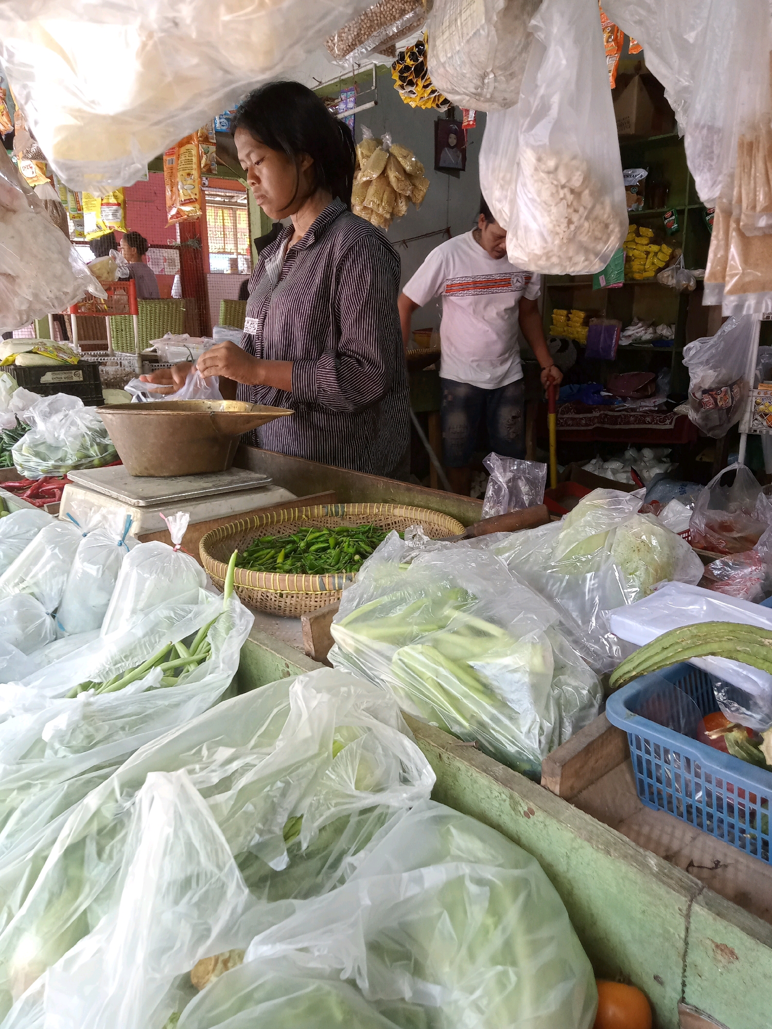 A woman stands at a market stall in Cirebon, Indonesia, preparing produce. She is wearing a striped shirt and appears to be weighing some items on a scale. The stall is filled with various fresh vegetables, some packaged in plastic bags, others in baskets. A man in a white t-shirt is visible in the background, also at the stall. The scene depicts a typical market environment with goods for sale and people engaged in transactions.