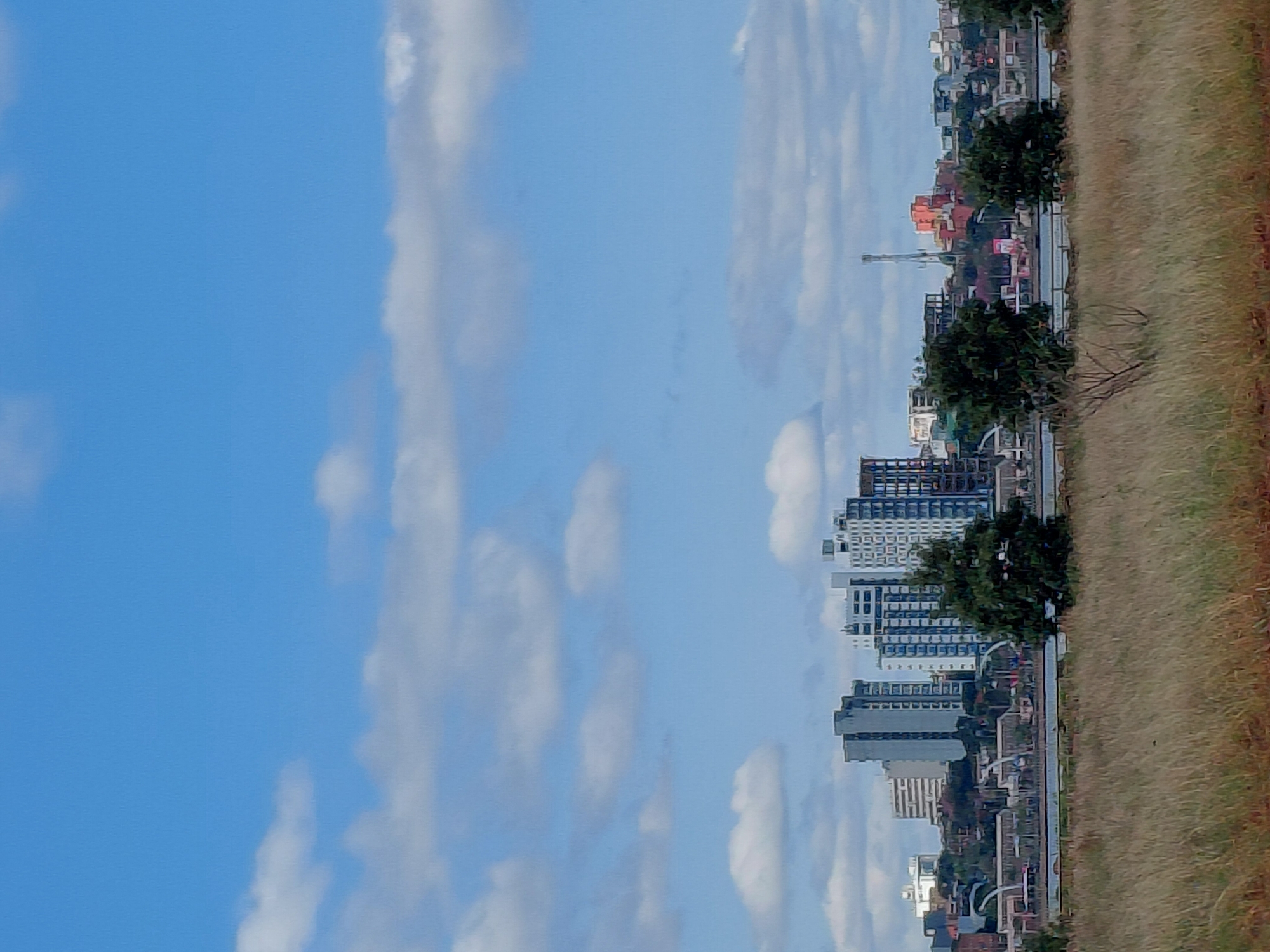 This image captures a panoramic view of the city of Encarnacion, Paraguay, from across a body of water. In the foreground, a field of dry, tall grass stretches towards the middle ground. Beyond the field, a calm body of water separates the viewer from the urban landscape.
The middle ground reveals a diverse cityscape. Several modern, multi-story buildings dominate the skyline, featuring a mix of architectural styles and colors. Some buildings are tall and slender, while others are wider and more block-like. White, grey, and blue facades are prominent. Interspersed among the buildings are areas of greenery, including trees. A distinctive communication tower with an orange structure is visible in the distance. Structures that appear to be bridges or elevated walkways can be seen spanning parts of the landscape.
The background is a clear blue sky filled with scattered white, fluffy clouds, suggesting fair weather. The overall impression is of a developed urban area situated by a waterfront, under a bright, partly cloudy sky. There are no visible people or animals in the image. The scene is tranquil and shows a cityscape with significant development.