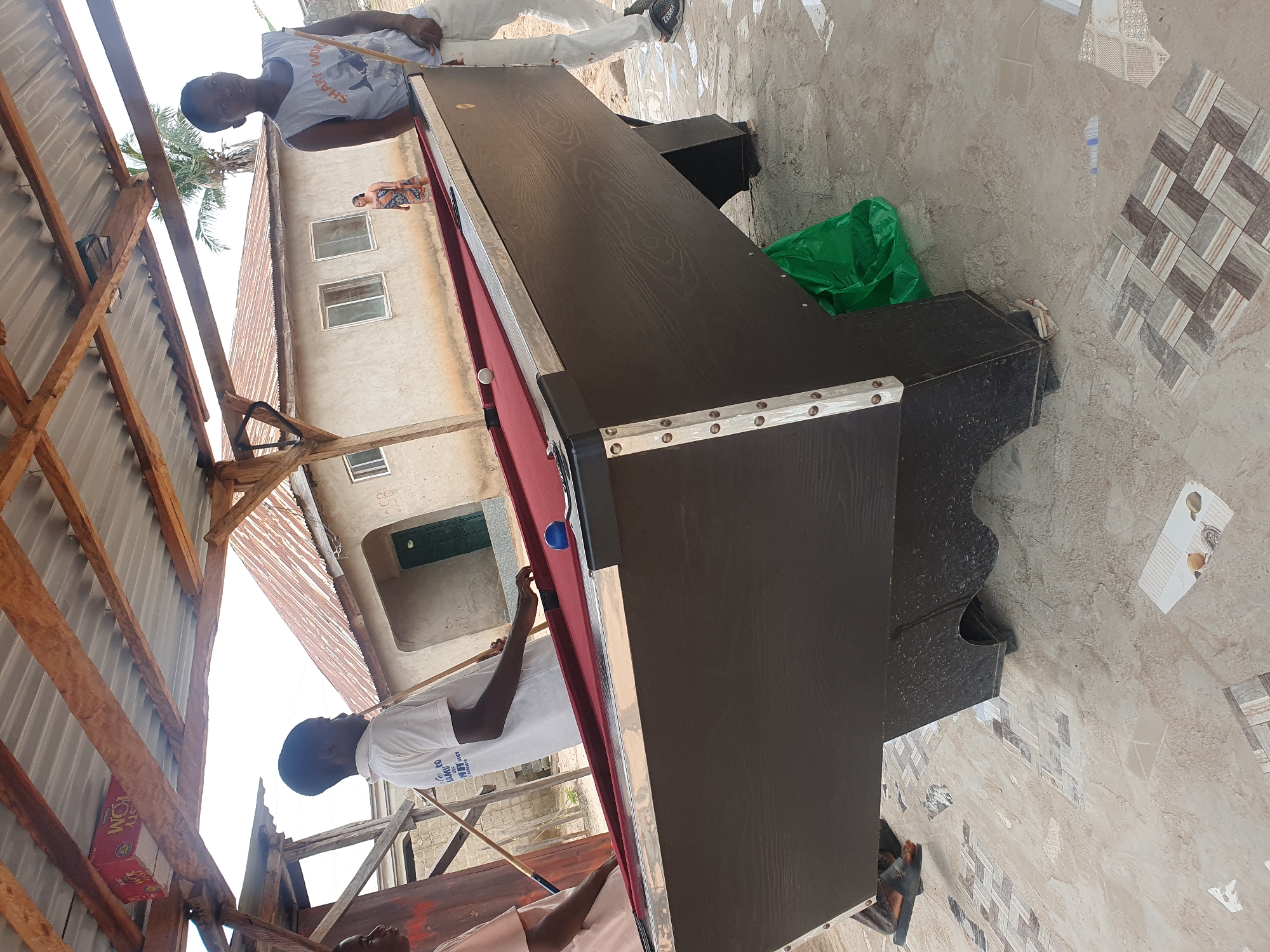 In Lokoja, Nigeria, beneath a corrugated tin roof supported by rough-hewn wooden beams, a pool table with a maroon felt surface is set up outdoors. Three young men are present. One, wearing a white t-shirt with text, stands on the left side of the table, holding a pool cue and seemingly focused on the game. Another young man, in a grey tank top and white pants, stands to the right, also holding a cue. A third person, partially visible on the far left, wears a light-colored garment. A blue pool ball rests on the table, and a white ball is visible further down. In the background, a building with a corrugated metal roof and closed doors and windows stands under a bright sky, with a palm tree visible behind it. The ground around the table is paved with stone tiles of various patterns. A green plastic bag is placed beneath the table.