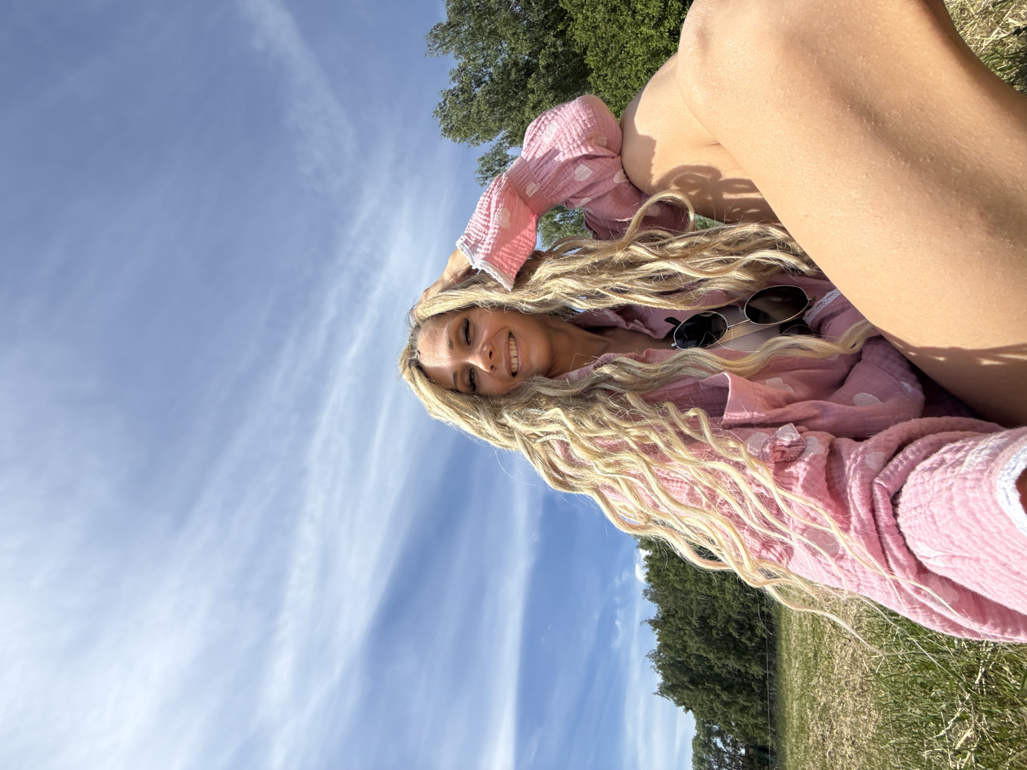 An outdoor scene in Rusanovo, Russia, depicts a woman seated on the ground in a grassy field under a bright blue sky with wispy white clouds. The woman, with very long, highly textured blonde hair (appearing as dreadlocks or similar braided style), smiles directly at the camera. She is wearing a loose-fitting, long-sleeved pink shirt with white circular patterns resembling polka dots or hearts, featuring white trim on the cuffs. Dark sunglasses hang from the neckline of her shirt. Her left hand is raised, with fingers lightly touching the side of her head. Her right leg is bent, with the upper thigh and knee prominently in the foreground. In the mid-ground, a line of dense green trees forms a natural backdrop. The lighting indicates a sunny day.