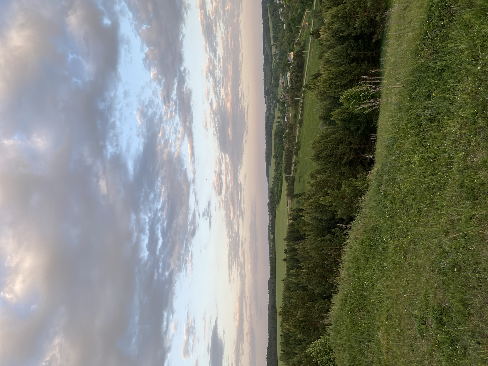 The image shows a landscape with a sky filled with clouds at sunset. Below the sky, there is a view of a field, a line of trees, and some buildings in the distance. The foreground consists of a grassy slope. The location is in the city of Unknown, Unknown.