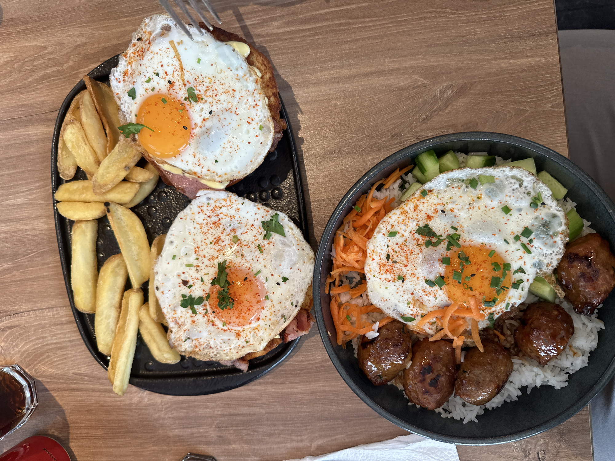 The image shows two plates of food on a wooden table. The plate on the left is black and contains two fried eggs on top of something, alongside potato wedges. A fork is visible, seemingly lifting a piece of the egg. The plate on the right is a dark grey bowl containing white rice, shredded carrots, cucumber slices, and what appears to be sausages, all topped with a fried egg. The eggs on both plates are sprinkled with herbs and spices. Parts of other items, like a small bottle and a piece of white cloth, are visible around the plates. The location and city of Sofia, Bulgaria is not evident in the image.