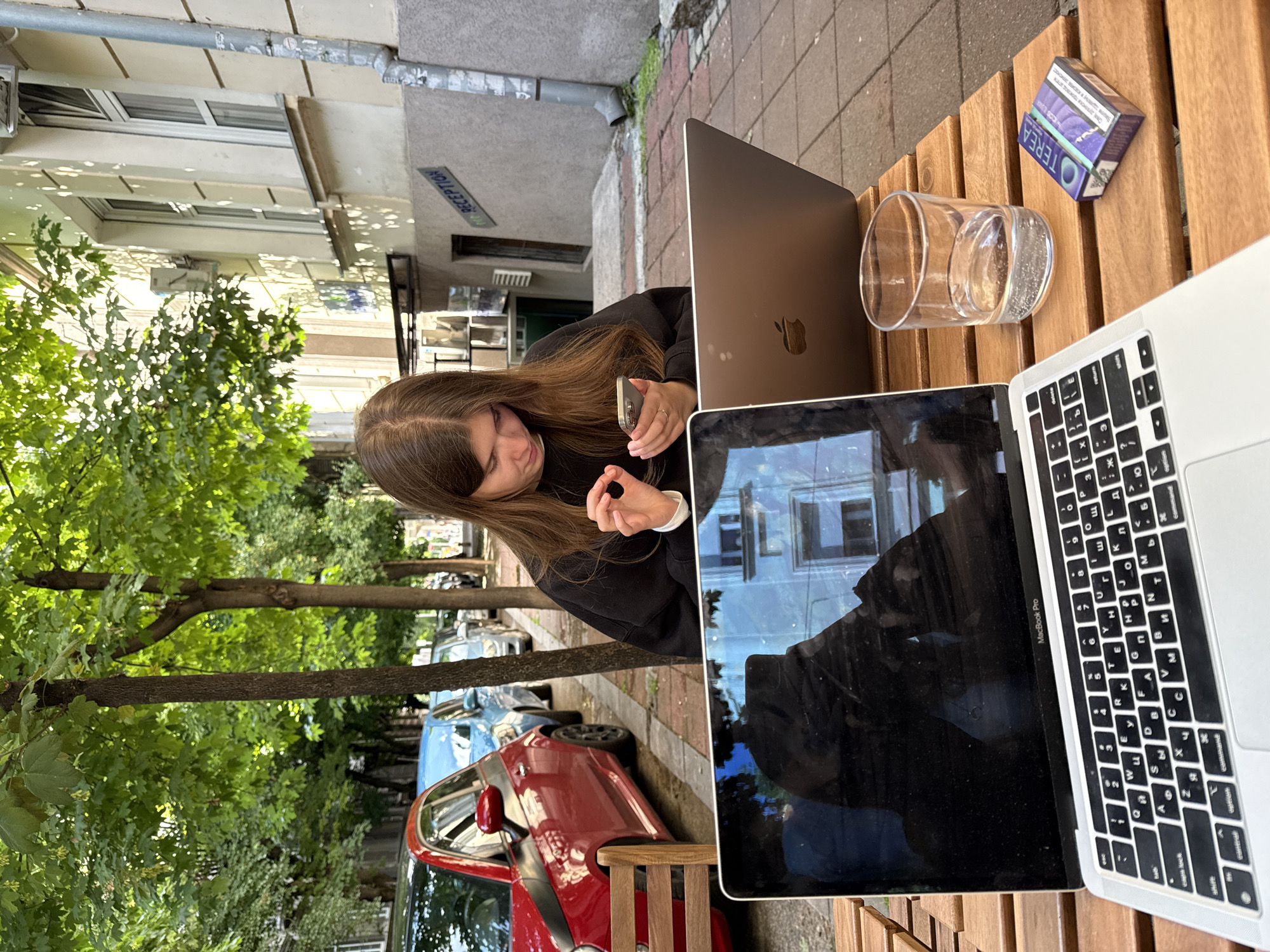 A medium shot captures a young woman seated outdoors at a wooden table in Belgrade, Serbia. She is holding a mobile phone in her left hand and a small black object in her right, with a focus on her action.
The table holds two open MacBook laptops, a glass of water, and a pack of Terea cigarettes. Behind her, the street scene includes trees, parked cars (one red and one blue), and a building with a sign reading "RECEPTION" in Cyrillic and English. The setting suggests an urban outdoor cafe or workspace.