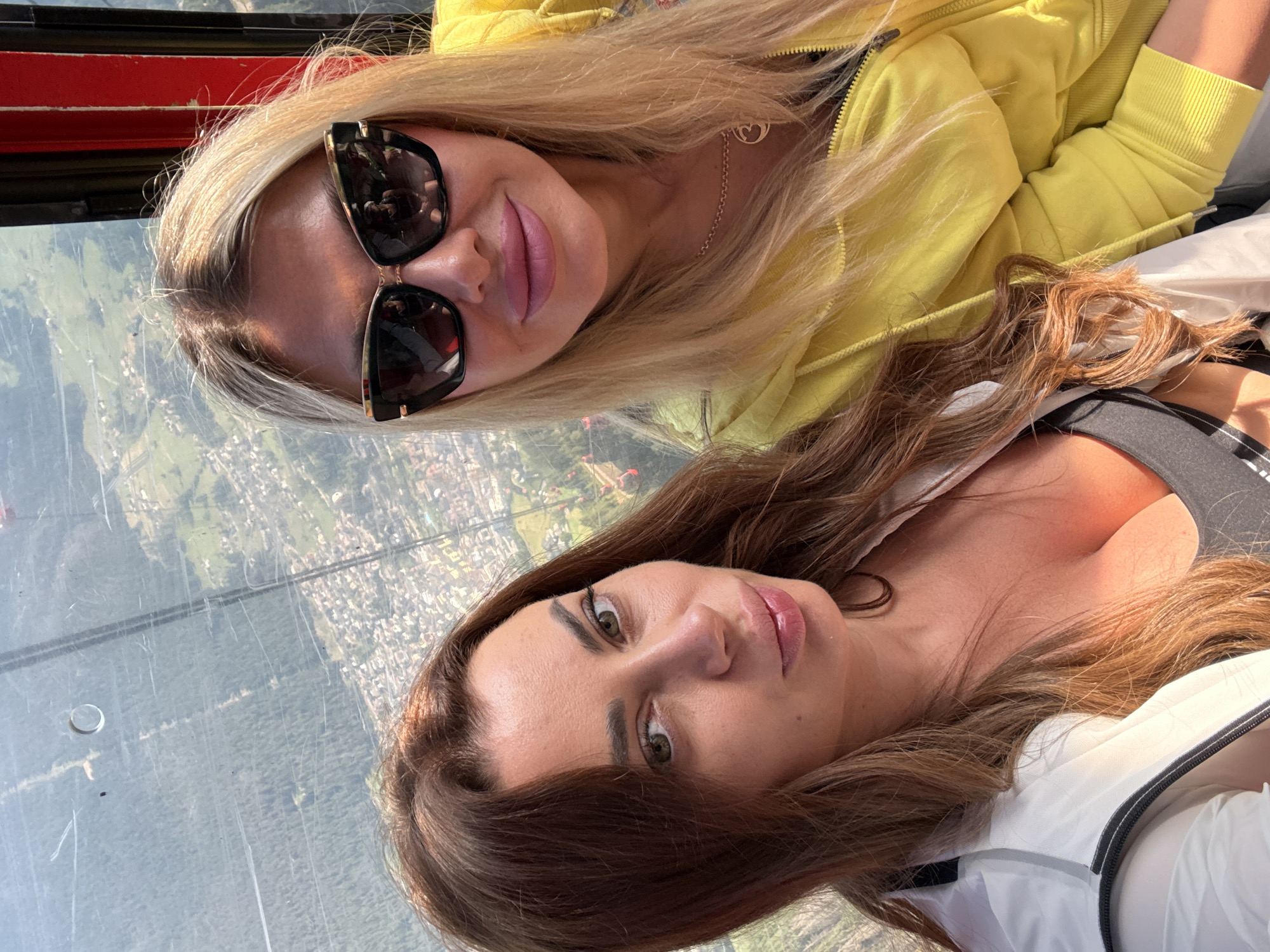Two women are taking a selfie in a cable car with a scenic mountainous landscape in the background. The woman on the left has long, wavy brown hair and is wearing a white jacket over a dark top. She is looking directly at the camera with light brown eyes. The woman on the right has long blonde hair and is wearing large, dark sunglasses and a bright yellow hooded jacket. She is smiling and looking at the camera. The cable car appears to be moving, as the background shows a village and a wooded mountainside. The exact location is not specified in the image but it is likely a tourist area.