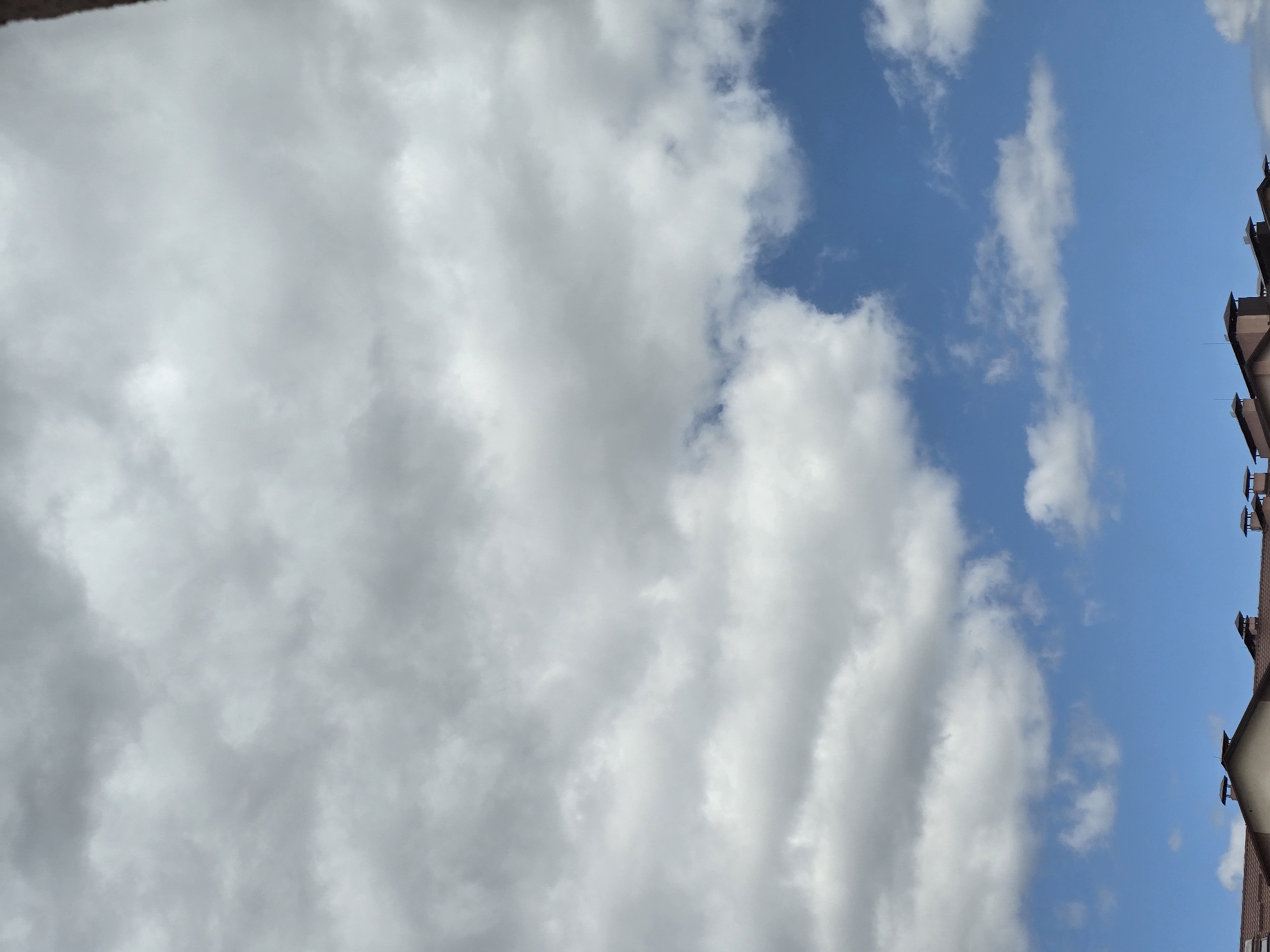 The image captures a view of the sky and rooftops in Sofiivska Borshchahivka, Ukraine. A predominantly cloudy sky fills the upper portion of the frame, with patches of bright blue visible. Cumulus clouds are present, varying in density and shade, suggesting a mix of sunlight and cloud cover. Below the sky, the brown-tiled roofs of buildings are visible along the bottom edge of the image. Several chimneys with dark caps protrude from these roofs. The architecture appears to be residential or commercial. The overall scene depicts an outdoor environment on what seems to be a partly cloudy day in an urban or suburban setting.
