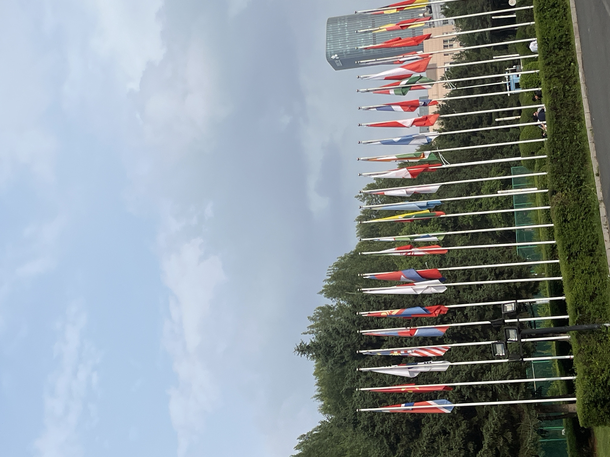 A line of flags of various nations flies against a cloudy sky in front of a dense line of evergreen trees. In the background, a modern, glass-fronted skyscraper stands tall. A low, manicured hedge runs along the foreground, with a paved surface visible at the very bottom of the frame. Several individuals are seated or standing near the hedge, some interacting with each other. The scene appears to be outdoors in a well-maintained urban area, possibly a park or plaza. The presence of numerous national flags suggests a diplomatic or international event is taking place in Changchun City, China.