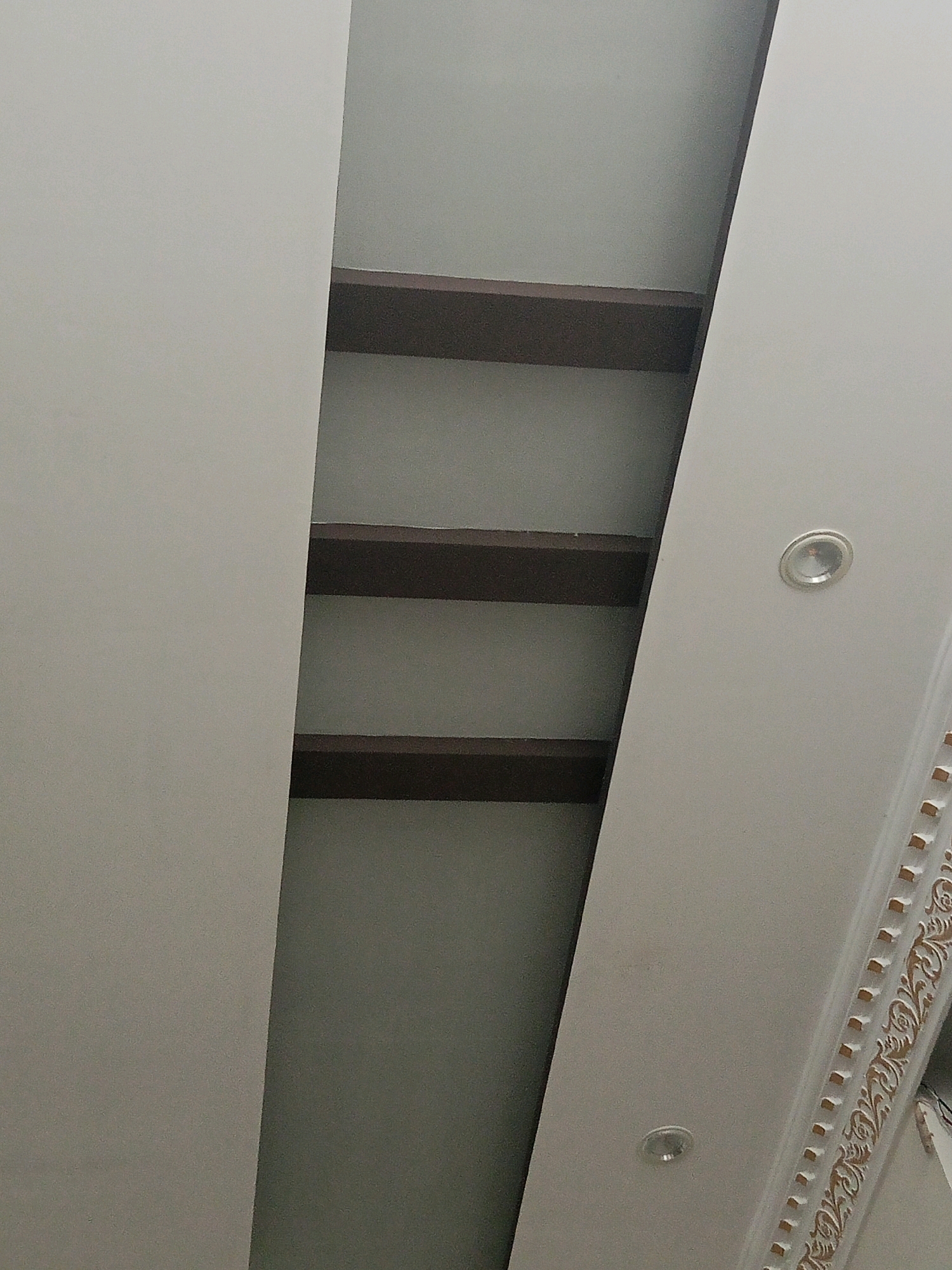 The image shows a view looking upwards at a ceiling in a building. The ceiling has a modern design with a recessed area that contains horizontal dark brown beams. There are also recessed circular lights embedded in the ceiling. The decorative trim along the edge of the ceiling is white with a gold floral pattern. The image does not contain any information about the city of Bhedian - Nai Basti, Pakistan or any people.