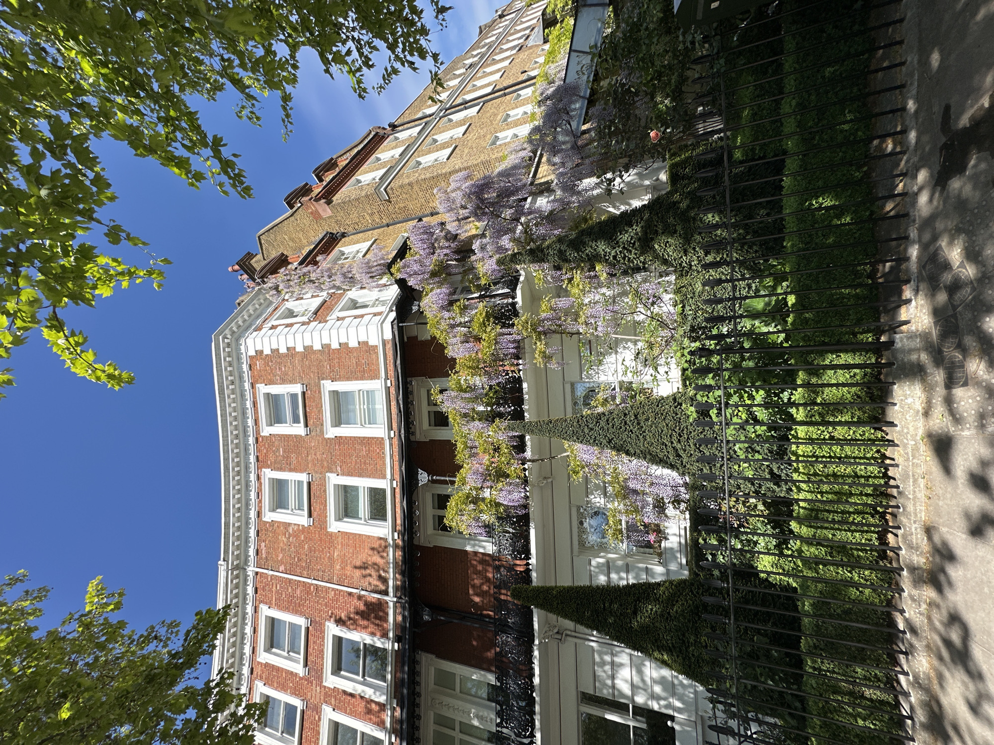A low-angle, outdoor photograph shows a multi-story building in what appears to be a wealthy neighborhood. The building, constructed primarily of brick and beige stone, is oriented vertically and slightly tilted, with a blue sky and sparse green foliage visible in the background. Wisteria vines with lavender-colored flowers cascade down the facade of the building. A wrought-iron fence runs parallel to the sidewalk, with ivy climbing along it. The sidewalk is made of gray stone pavers. There are no people visible in the image. The location appears to be a residential area of London, United Kingdom.