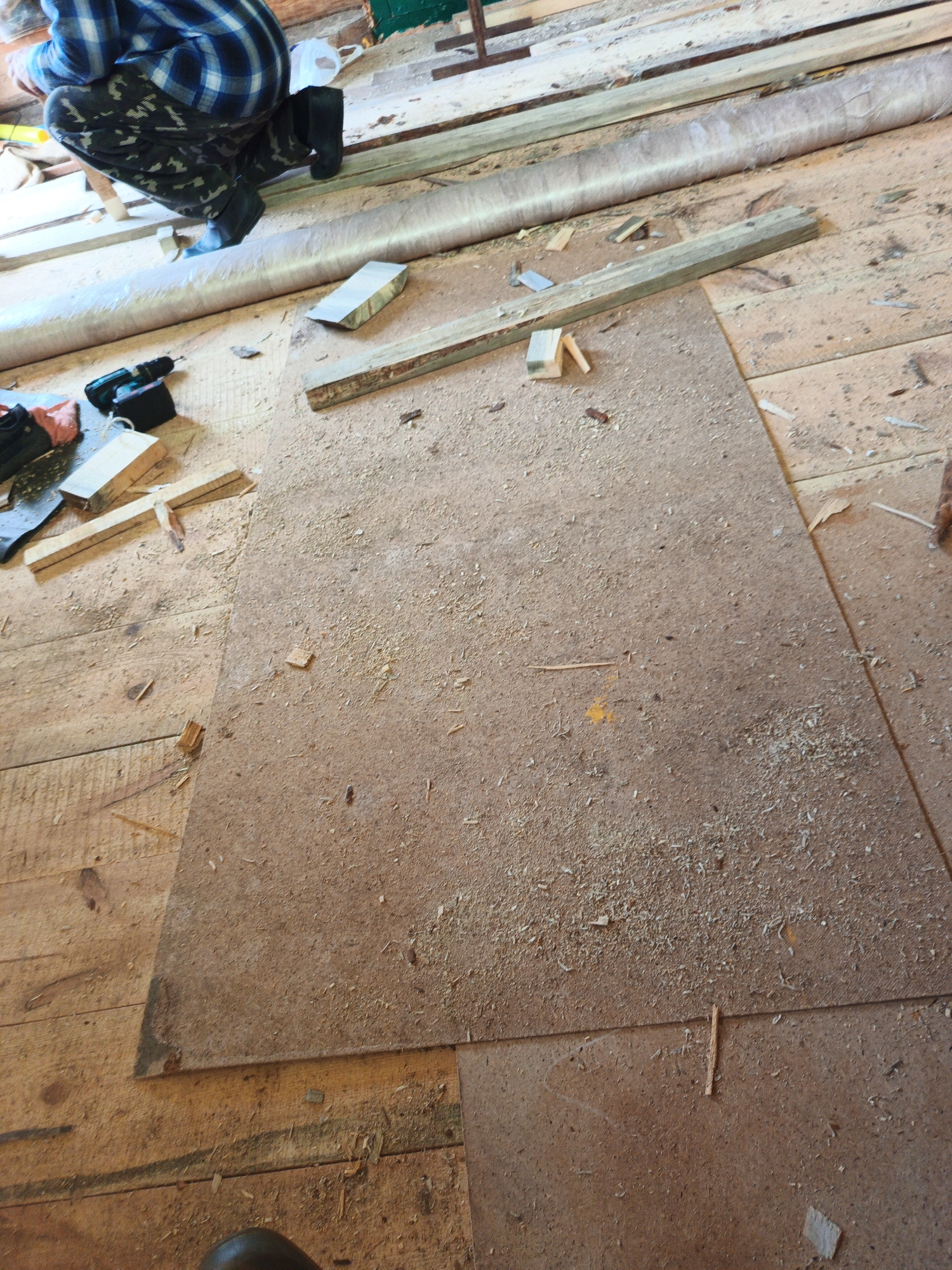 In Islambaevo, Russia, a person wearing a blue and black plaid shirt, camouflage pants, and black boots kneels on a wooden floor covered with sawdust and scattered wood scraps. The person is near a workbench area where a cordless drill, tools, and pieces of wood are visible. A large piece of pressed wood or particle board lies on the floor in front of the person, also covered in sawdust. The environment appears to be an indoor workshop or construction site, with unfinished wooden planks forming the floor and walls. The scene suggests ongoing construction or renovation work.