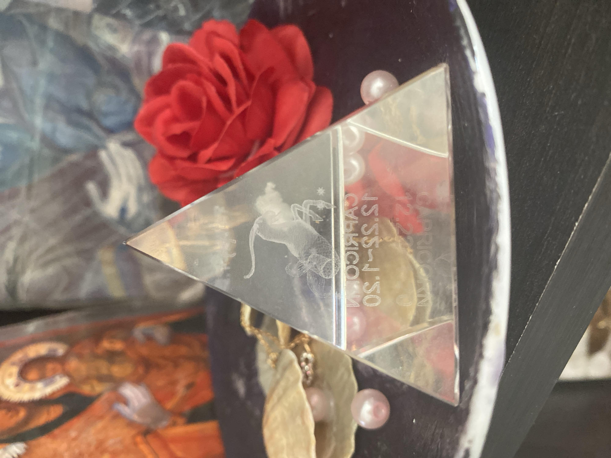The image displays a still life arrangement on a dark surface, likely a shelf. The central focus is a clear glass pyramid, etched with the Capricorn zodiac symbol of a goat. The pyramid is positioned on a dark, possibly velvet, circular mat. To the left of the pyramid, a seashell holds several pink pearls and a gold necklace. Another single pink pearl rests on the mat in front of the seashell. Behind the pyramid and mat, a bright red artificial rose is visible, and further in the background, out of focus, are what appear to be religious icons or paintings, one featuring figures in orange and gold robes. The overall scene is dimly lit, with soft light illuminating the objects. There are no people present in the image. The location in Blagoevgrad, Bulgaria, is not discernible from the visual information provided.