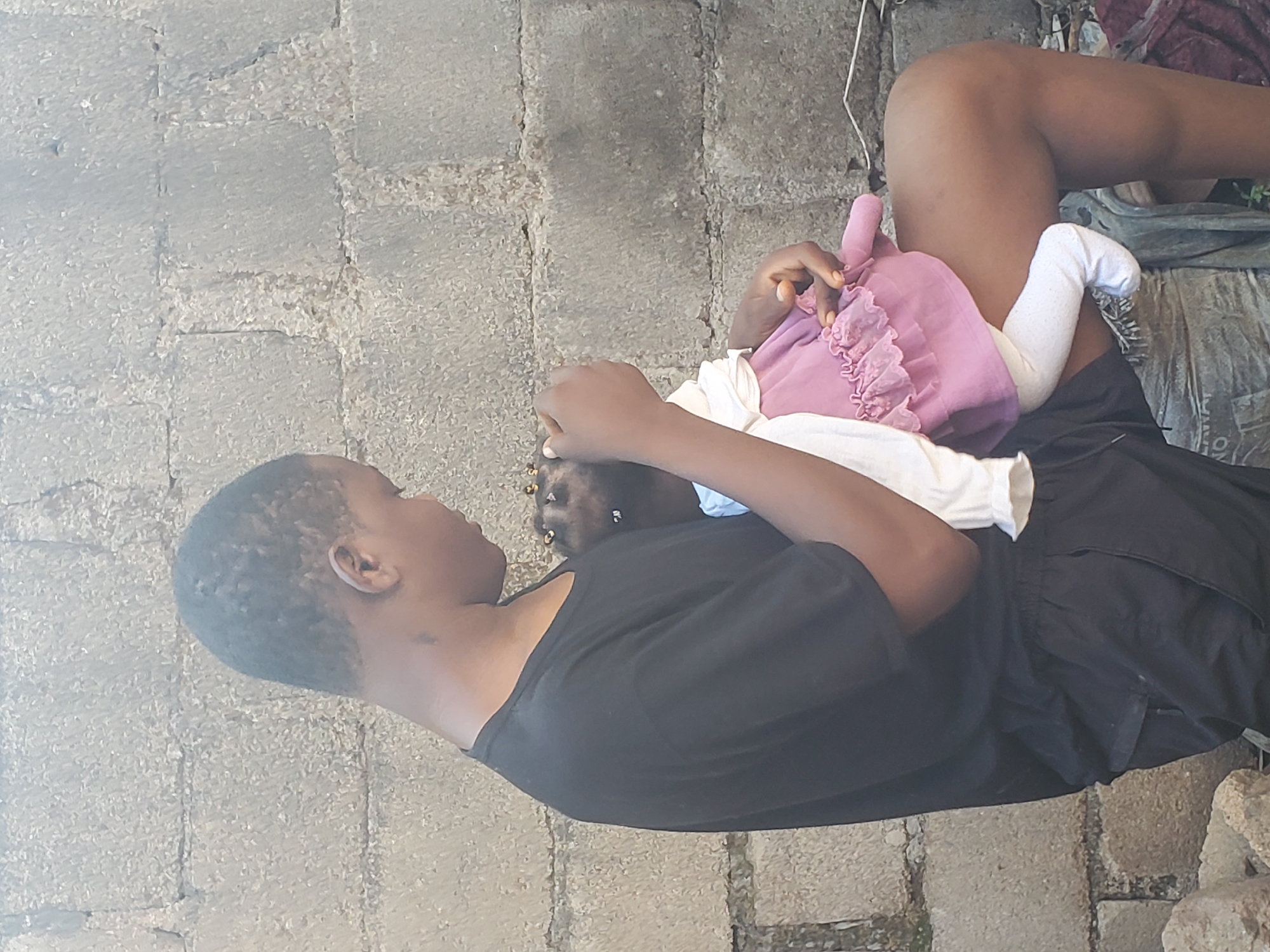 The image displays an adult and an infant in an outdoor setting. An adult, with dark skin and short dark hair, is lying on their back, wearing a black t-shirt and black shorts or trousers. They are holding an infant who is resting on their stomach or chest. The infant is dressed in a light purple ruffled garment over a white top, with white socks or leg coverings on their feet. The adult's head is turned towards the infant, suggesting a tender and attentive moment.
The background consists of a textured, grey paved surface, likely made of concrete or stone blocks, which appears to be ground cover. The paving shows cracks and irregularities. In the lower right section of the image, there are some rough stones and what appears to be discarded debris or fabric. The lighting indicates it is daytime, with soft, diffused illumination. There is no visible text in the image. The scene conveys a quiet, intimate moment of care between the adult and the baby in a casual outdoor environment in Jos, Nigeria.