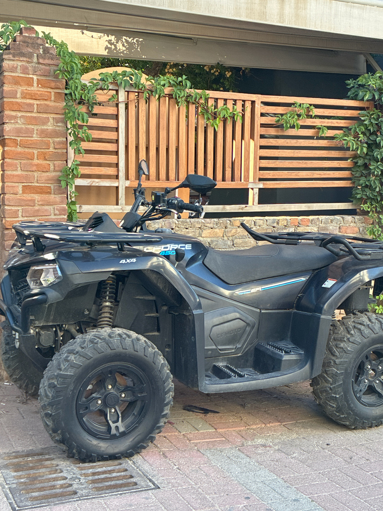A dark gray CFMOTO 450 4x4 ATV is parked on a brick-paved area in Alanya, Turkey. The ATV is positioned in front of a wooden slatted fence and a brick wall. Green ivy vines are growing on the fence and the brick wall. The ATV has a black seat, black tires with visible tread, and black racks on the front and rear. The CFMOTO logo is visible on the side panel. The lighting suggests it is daytime, with sunlight casting shadows. There are no people visible in the image.