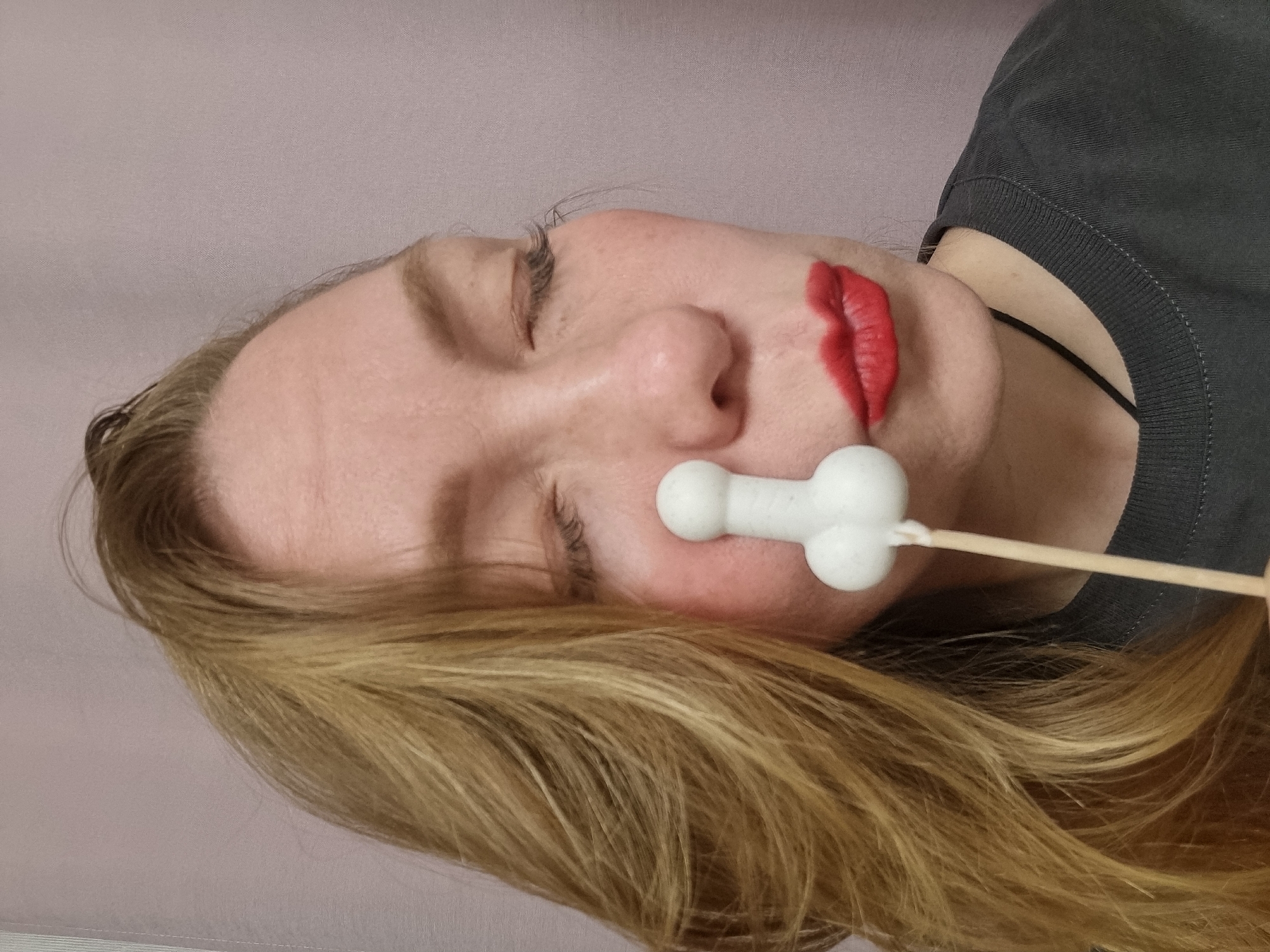 A woman with blonde hair is lying down with her eyes closed. She has red lipstick on. A white, bone-shaped object on a stick is held near her nose. The woman is wearing a gray shirt. The background is a light pink. There is no evidence of the location being in Chaikovsky, Russia.