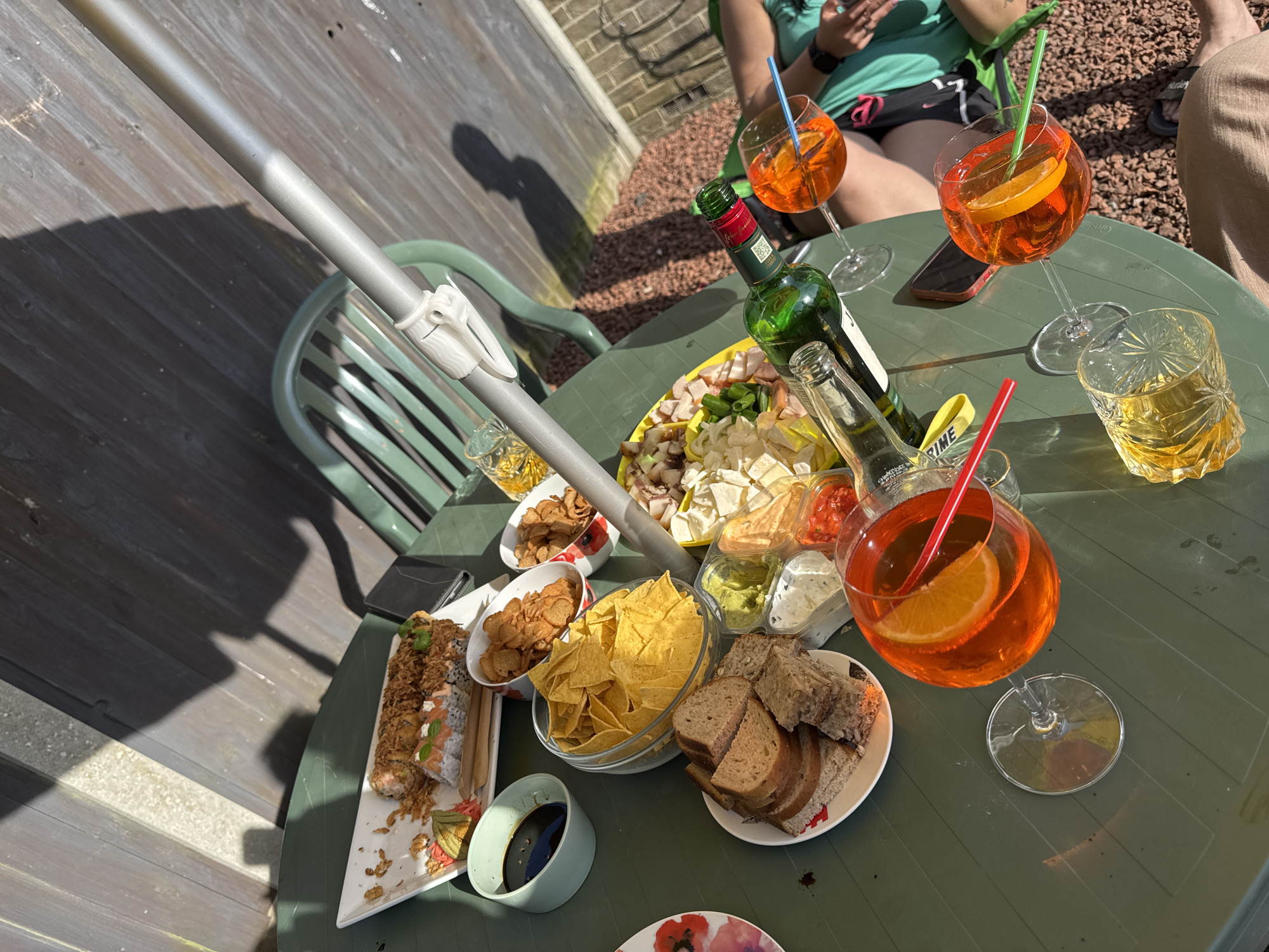 The image shows a green outdoor table covered with various food items and drinks. The food includes sushi rolls, chips, bread, and a platter of cheese and meats with dips. Drinks include Aperol spritzes in large glasses, and an unidentified amber beverage in a decorative glass. There are two people present, partially visible. One person is wearing a green top and black shorts, looking at their phone. The second person is wearing tan pants. The setting appears to be a backyard or patio, with a wooden fence and paving stones in the background. The scene is sunlit, with shadows cast by an umbrella pole. There is no evidence to support the location being Great Yarmouth, United Kingdom.
