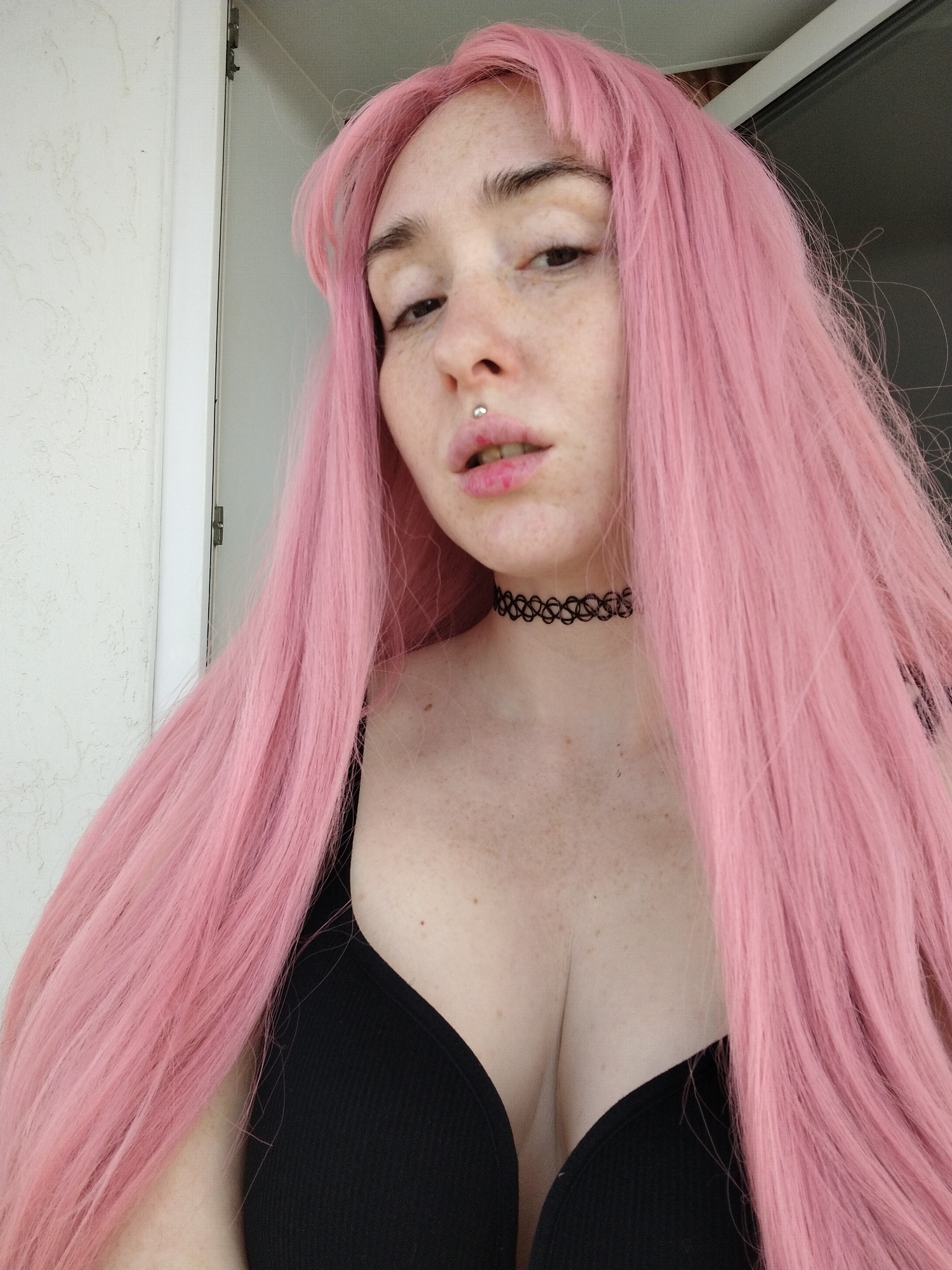 A woman with pale skin and freckles is posing for a photograph. She has long, pink hair with bangs and is wearing a black choker necklace and a black tank top. There is a lip ring on her upper lip. The background shows a white wall and part of a doorframe. There is no indication the image was taken in Izhevsk, Russia.