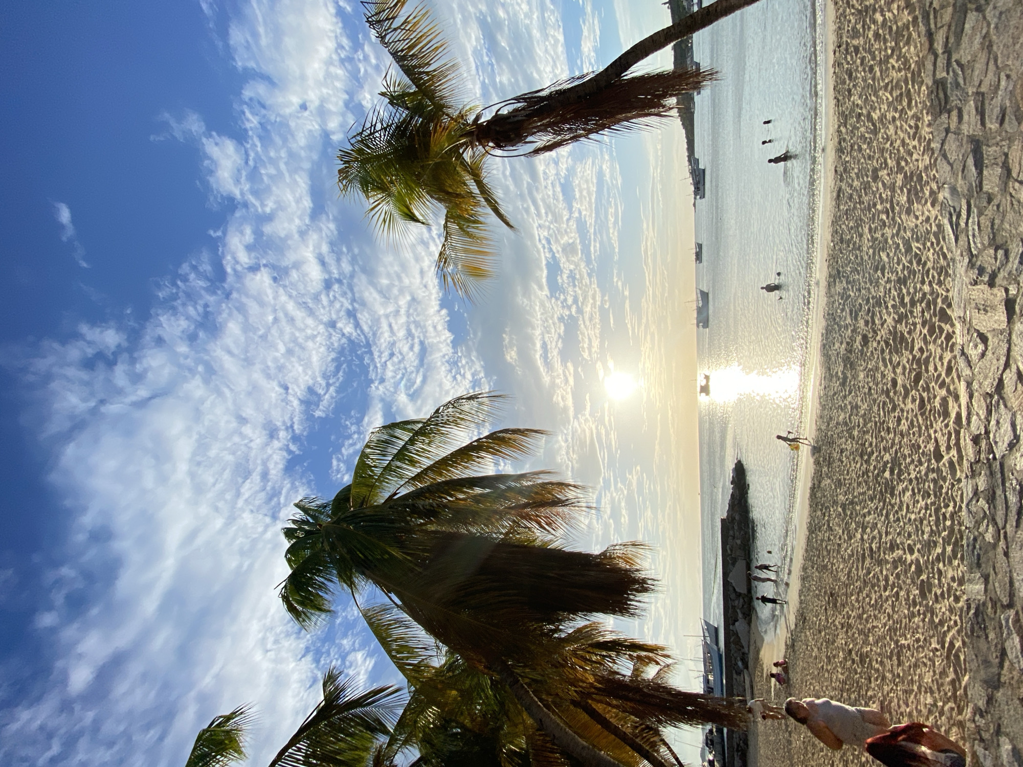 This is a photograph of a beach scene in Juan Griego, Venezuela, featuring palm trees, the ocean, and several boats. The sky is a bright blue with scattered white clouds, and the sun is setting or rising, casting a warm glow over the water. Several people are visible on the beach and in the shallow water. Some are walking along the sand, while others appear to be swimming or playing. A few boats are anchored in the distance, and the shoreline is lined with sand and some rocky structures. The palm trees are positioned in the foreground and midground, with their fronds extending into the frame and partially obscuring the sky. The overall atmosphere is serene and summery.