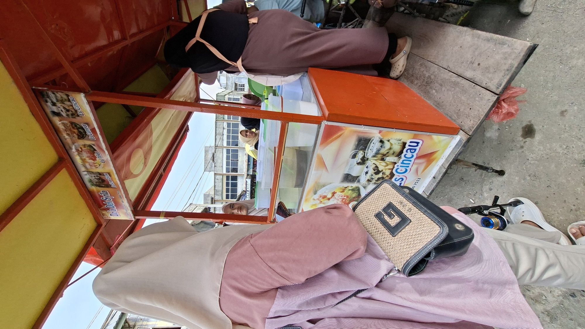 In this media file, the scene captures an outdoor setting with a focus on a food cart or stall. Two individuals are partially visible, one wearing a pink headscarf and pink striped shirt, and another wearing a black top and a muted pink skirt or dress. The food cart is brightly colored in red and yellow, featuring a display of drinks and food items. Visible on the cart are signs with text in Indonesian, including "NAVA KIDS," "Sirup Cincau," "Es Teler," "Es Campur," and "Es Cincau." A woven handbag with a prominent "G" logo sits on the lap of the person in the foreground. In the background, buildings with windows and a glimpse of the sky suggest an urban or semi-urban environment. The ground appears to be paved or concrete. The lighting suggests it is daytime, and the overall atmosphere seems to be casual and possibly commercial, with the food cart indicating a point of sale for refreshments.