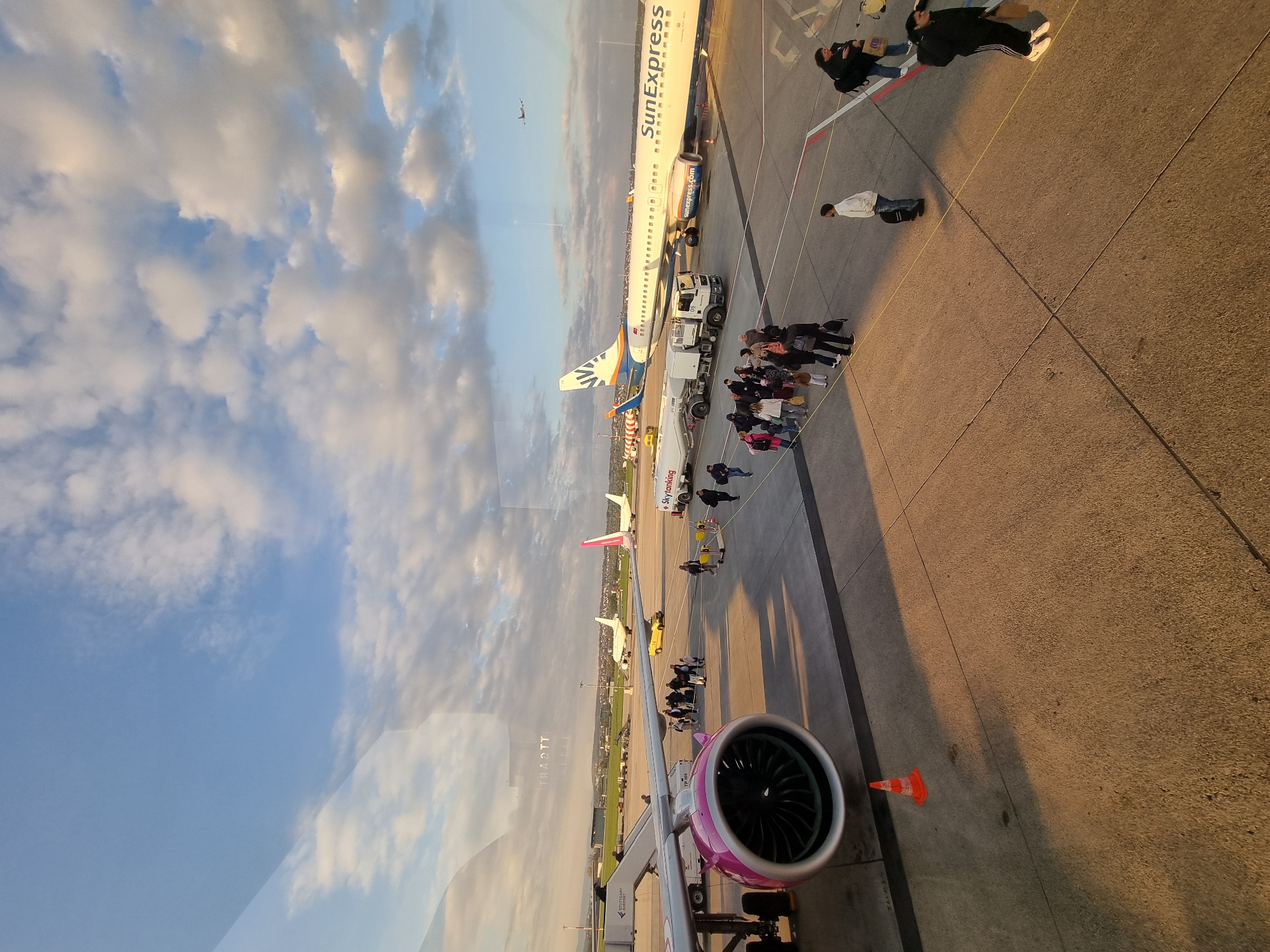 The image is taken from inside an aircraft or airport terminal, looking out onto an airport apron in Echterdingen, Germany. The sky is partially covered with clouds. A SunExpress aircraft is parked on the apron, and passengers are boarding or disembarking. There are several other aircraft parked in the background, including one with a pink tail. A jet bridge with a pink engine is visible in the foreground, presumably connected to the aircraft where the photo was taken. Airport ground support vehicles are visible near the SunExpress plane. Groups of people are standing around, likely waiting to board or having disembarked from a plane. An orange cone is near the engine.