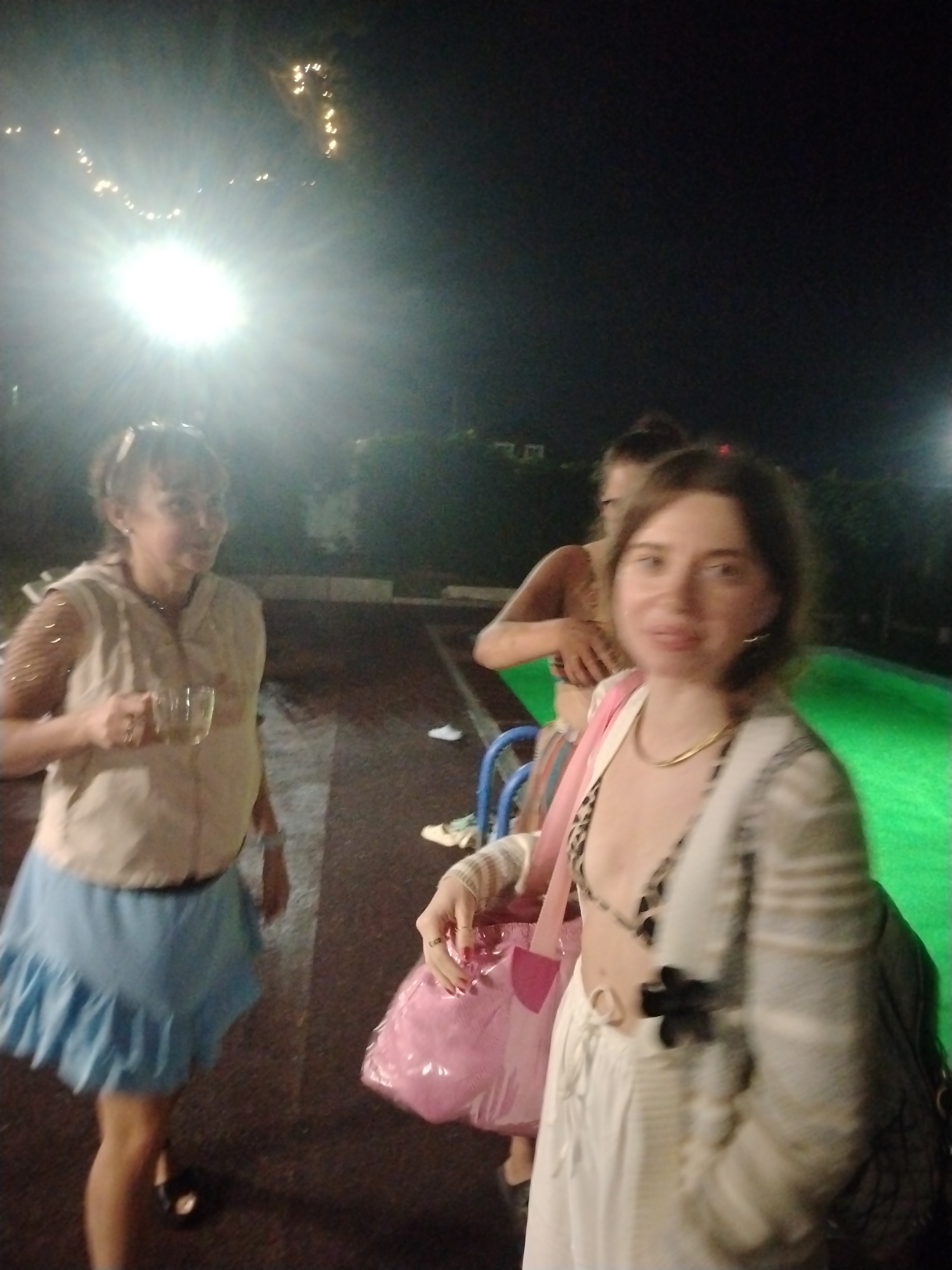 The image shows three people outdoors at night, with a bright light source in the upper left corner illuminating the scene. In the foreground to the left, a woman with brown hair tied up is wearing a beige sleeveless vest over a blue ruffled skirt. She is holding a clear glass with a liquid. To the right of her, another woman with brown hair is wearing a bikini top with a leopard print, a white skirt, and a striped cardigan. She is holding a pink bag and has a gold necklace. Behind them, partially visible, is a third person, also with brown hair, wearing a bikini top. The background shows some trees and what appears to be a paved area. The lighting suggests this could be a public space like a park or an event area. The presence of string lights in the background further hints at a festive or recreational setting. Without additional context or identifiable landmarks, it is not possible to definitively determine the location within Chaikovsky, Russia.