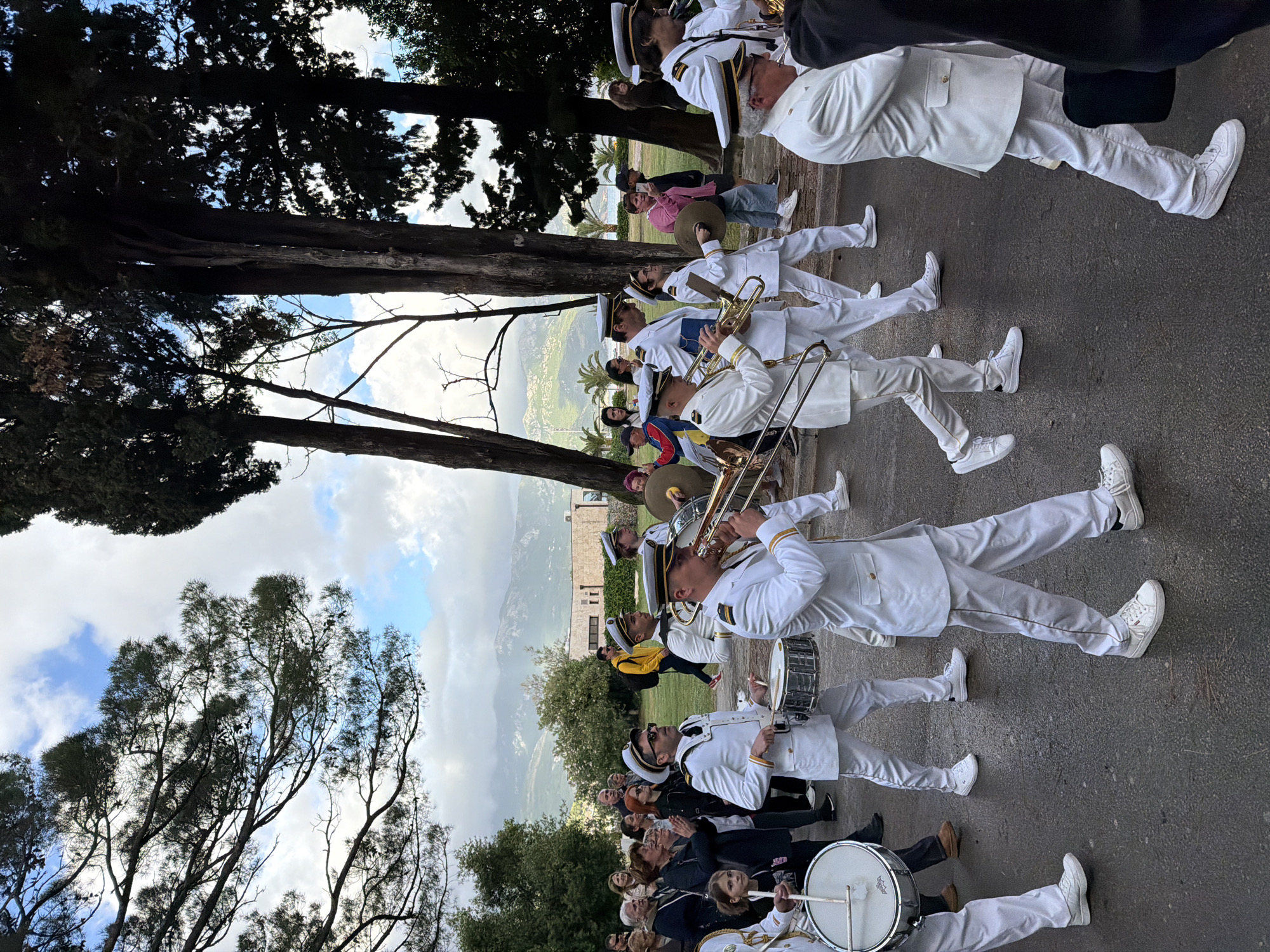 A horizontal, medium-shot image shows a brass band dressed in white uniforms, moving from the right side to the left. The band marches along a road lined with people watching, on the right, and trees on the left, under a partially cloudy sky. In the background, a view of mountains and buildings can be seen. The photo was taken outdoors in Budva, Montenegro.