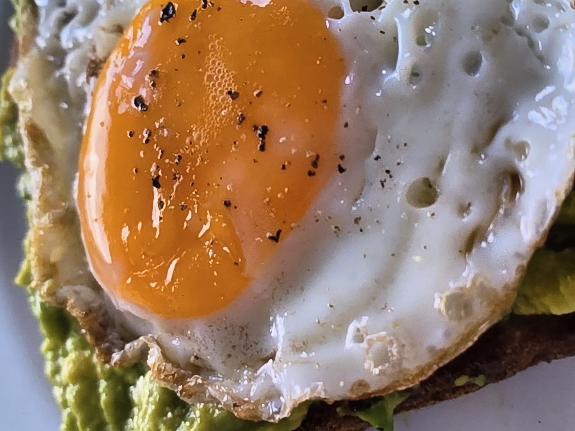 A close-up, high-angle shot shows an open-faced sandwich with mashed avocado and a fried egg. The sandwich is on a slice of toast, and the avocado is spread evenly on top of the toast, followed by a sunny-side-up fried egg. The egg has a bright orange yolk and a white cooked albumen. Black pepper is sprinkled over the yolk. The background is out of focus, with a hint of white plate visible. This image was taken in a home kitchen in Voluntari, Romania.