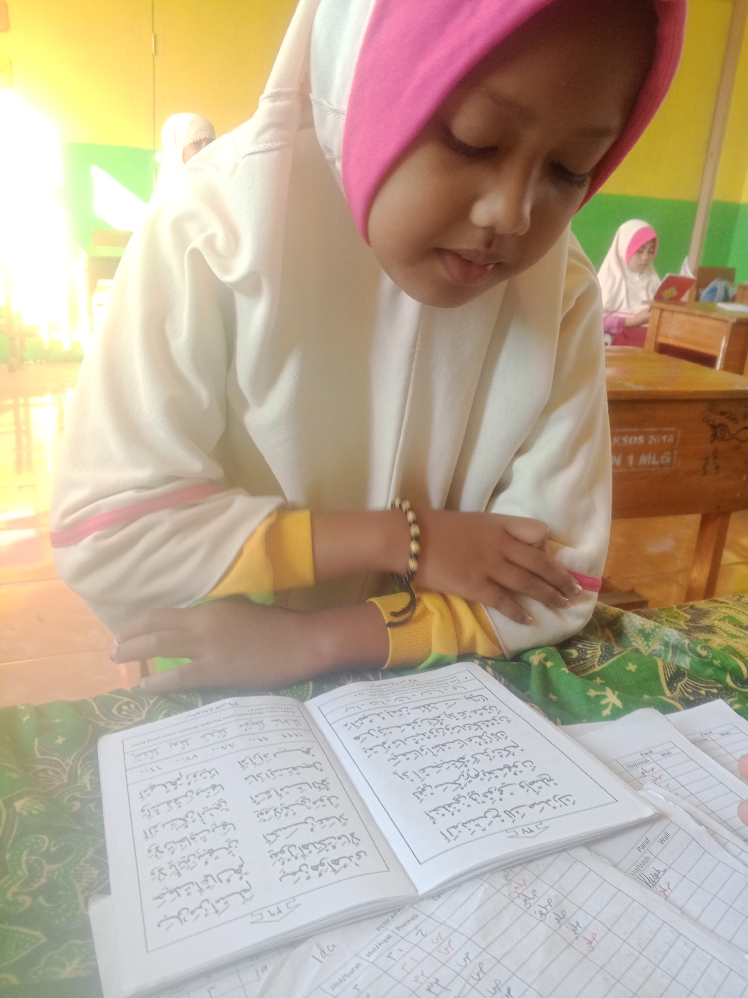 A young girl wearing a pink and white hijab and a cream-colored outfit is sitting at a desk, looking down at an open book with Arabic text. She is in the foreground, with her hands resting on the book. Another girl, also wearing a hijab, is visible in the background, sitting at a desk. The environment appears to be a classroom, with wooden desks and a yellow wall. The desks have inscriptions on them, one reading "KSOS 2018" and "N 1 MLG". The image was taken in Kemulan, Indonesia.