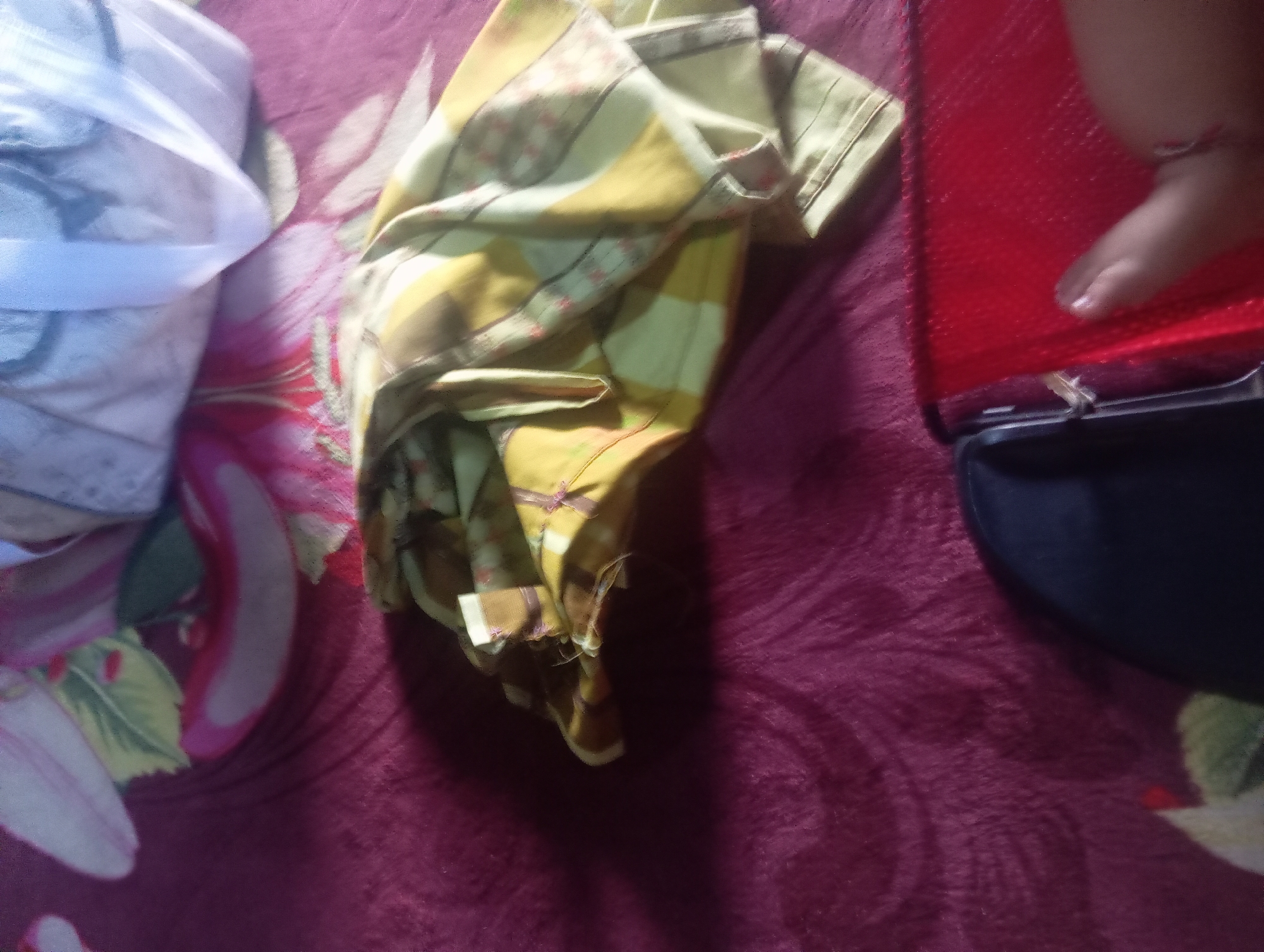 In Rejosari, Indonesia, a close-up, overhead shot displays a crumpled, light yellow and green patterned garment, possibly a piece of clothing, resting on a deep purple, textured surface that appears to be a blanket or rug. To the left, a white, translucent fabric is visible, partially covering a pink and green floral pattern on the purple surface. To the right, a red, textured object, possibly a book or binder, is partially obscured by a hand with a light brown fingernail polish. A dark object, potentially a phone or other device, is positioned beneath the red object. The lighting suggests indoor illumination, casting shadows across the scene.