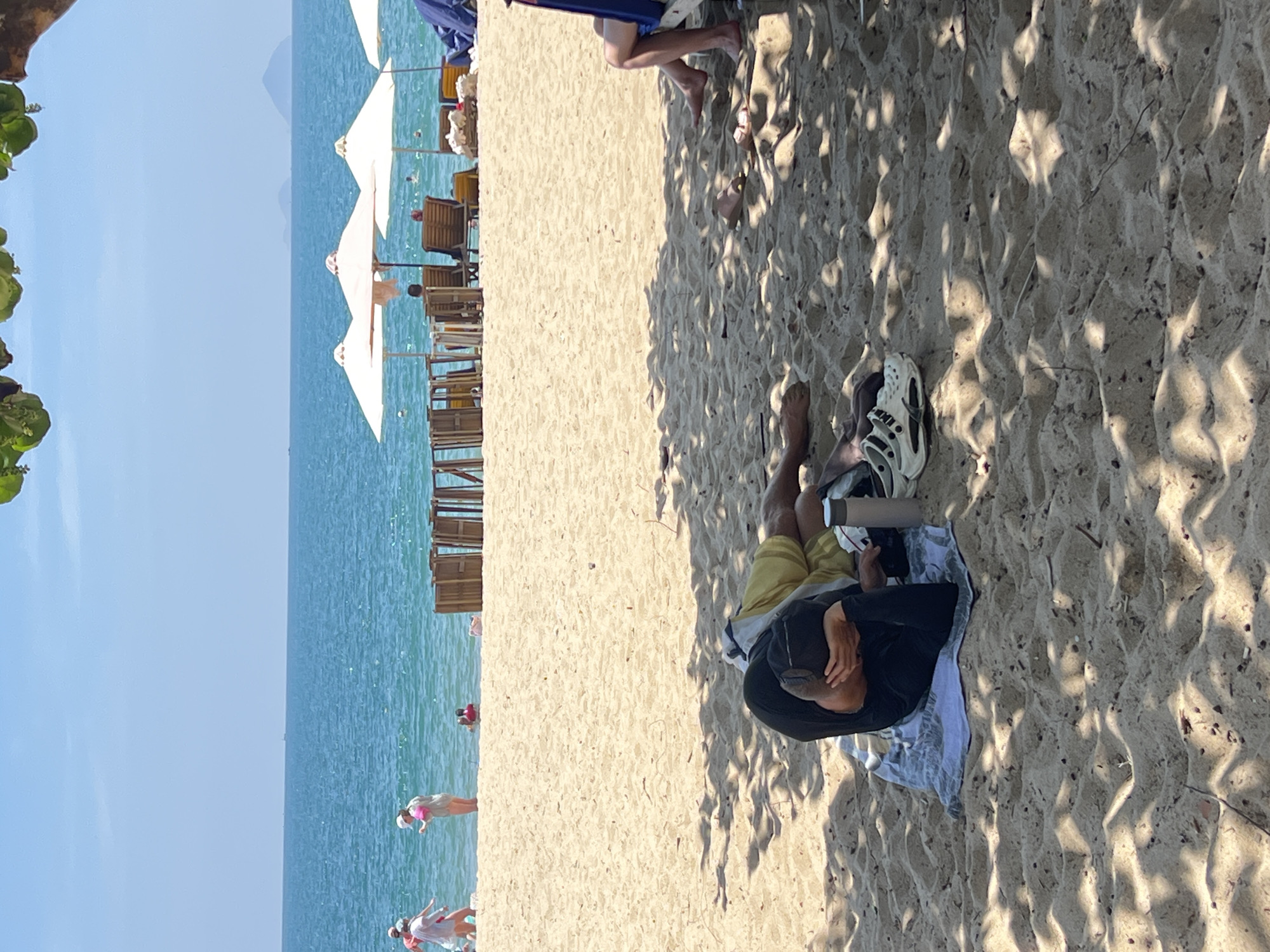 A person is lying prone on a light-colored sandy beach, partially shaded by foliage. They are wearing a black shirt and yellow shorts, and their head is turned towards their right side. Adjacent to the person is a water bottle. A beach towel partially covers them, and nearby there are two shoes and feet. Beyond the beach, the ocean stretches to the horizon, with several white beach umbrellas and chairs positioned near the water's edge. The sky is a clear blue. People are wading in the water, and the location appears to be a beach near Nha Trang Ward, Vietnam.