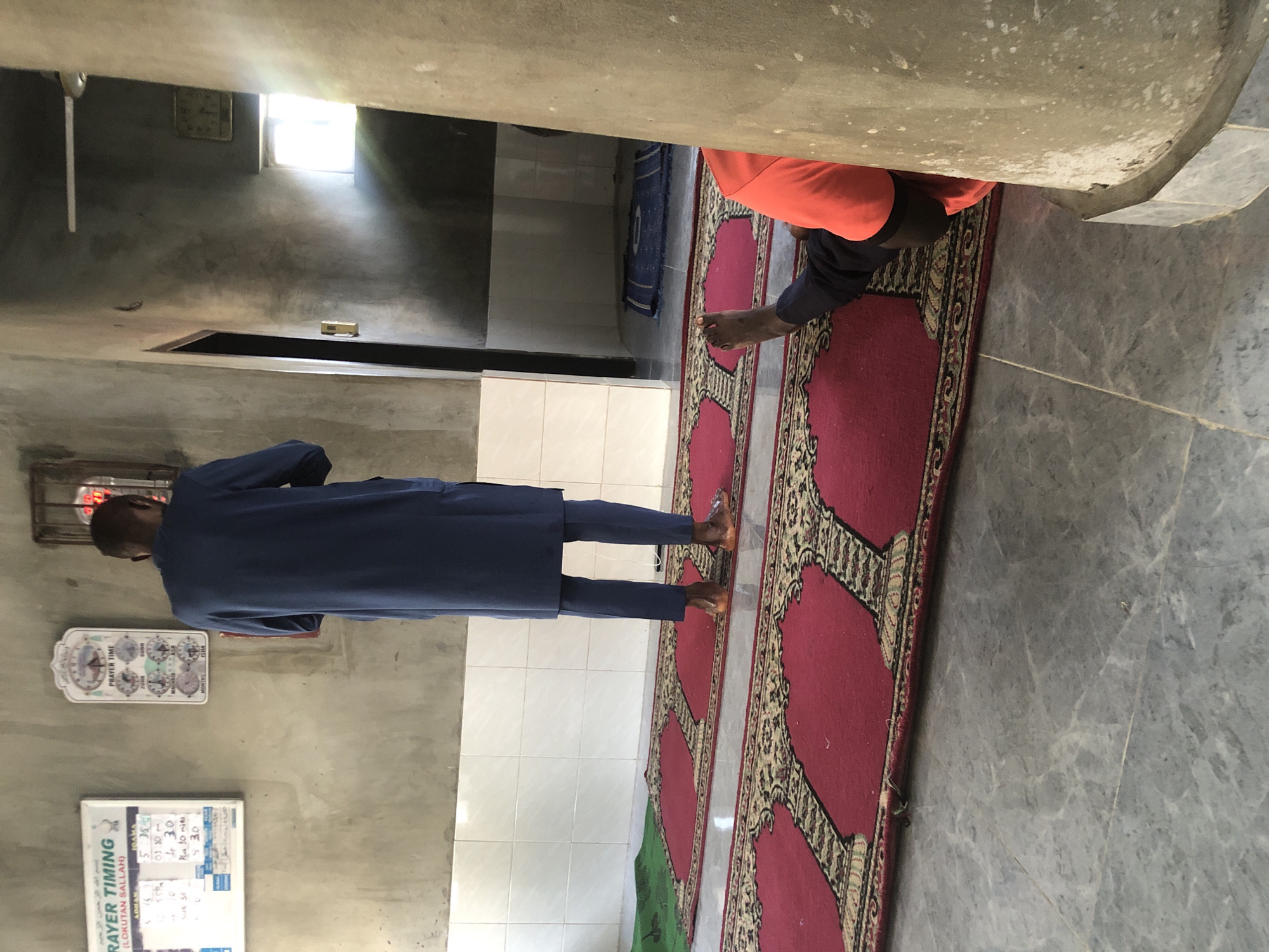 The image depicts the interior of a mosque, likely in Ado, Nigeria. Two individuals are visible. One man, wearing a blue tunic and trousers, stands facing away from the camera, appearing to be in prayer. Adjacent to him, on the floor, is another person, partially visible, wearing an orange shirt and dark trousers, with their legs outstretched. Red prayer rugs are laid out on the floor. The walls are concrete, with a section of white tiles above the prayer rugs. A prayer time chart is mounted on the wall to the left of the standing man. A ceiling fan is visible towards the back of the room, and a window lets in natural light. The floor is tiled with a gray marbled pattern.