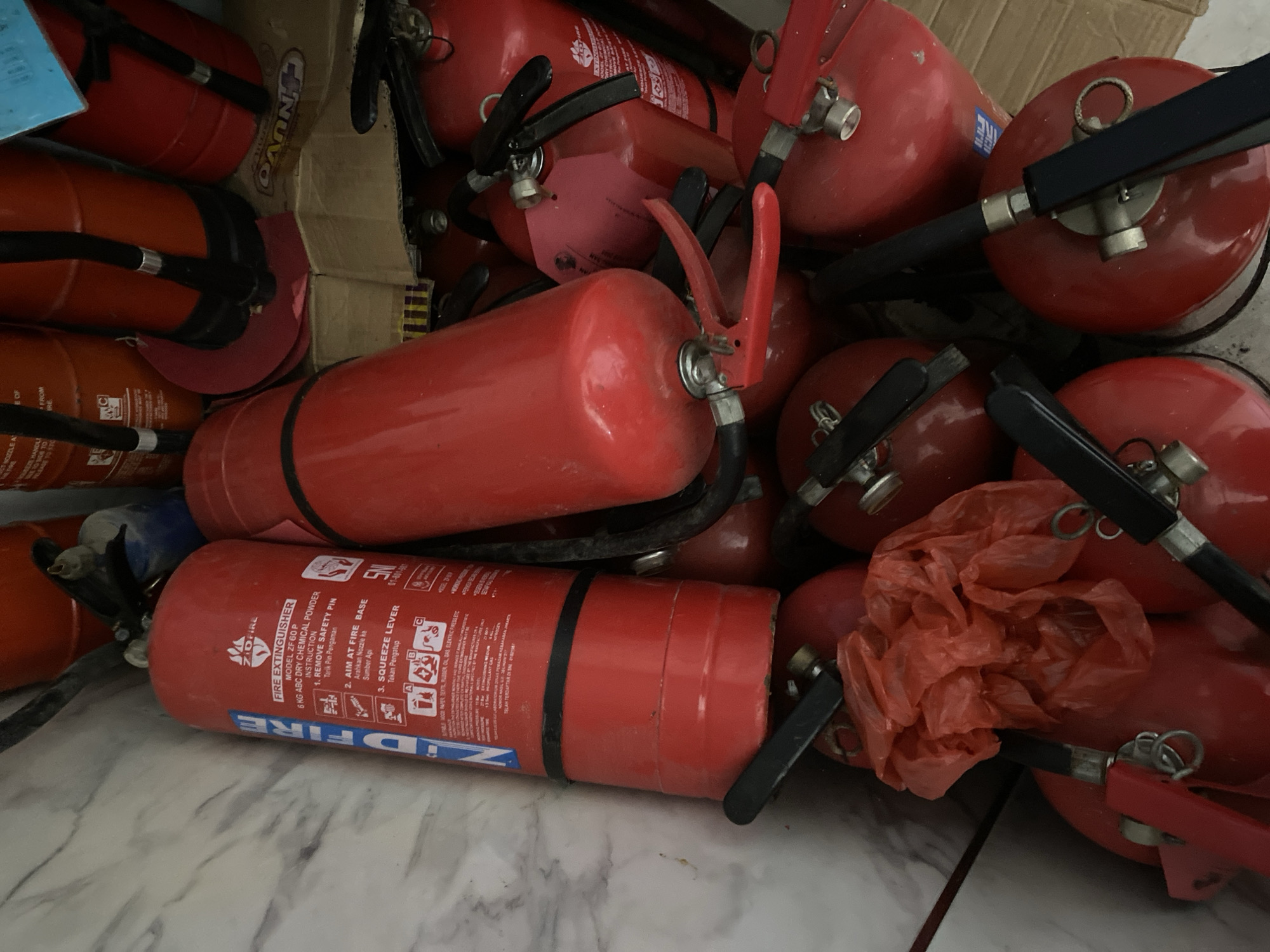 A high-angle, close-up shot shows a collection of red fire extinguishers piled together in a storage area. The fire extinguishers are of various sizes, with some larger units featuring black hoses and red handles, and smaller units with black handles. Labels and instructions are visible on some of the larger extinguishers. One fire extinguisher in the foreground has a "ZIDFIRE" label and displays instructions for use. A crumpled red plastic bag is visible amongst the extinguishers. Cardboard boxes are partially visible in the background. The environment appears to be a dimly lit indoor space, possibly a storage room or warehouse.