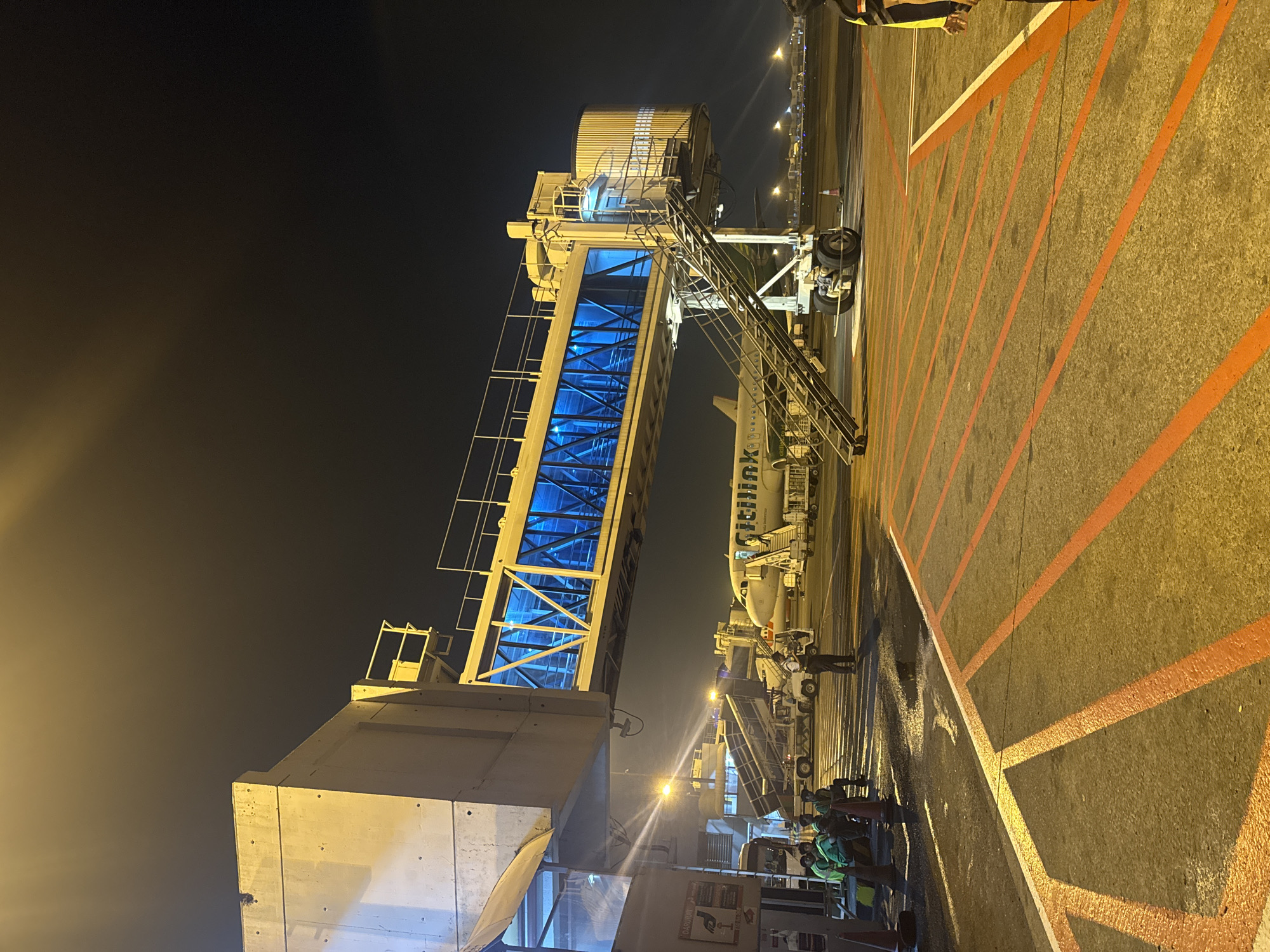 The image shows a nighttime scene at Soekarno-Hatta International Airport (CGK) in Tangerang, Indonesia. An airport jet bridge is connected to a Citilink Airbus A320 aircraft. Several airport ground crew members are visible near the aircraft and jet bridge, some appearing to be involved in loading or unloading activities. The tarmac is marked with orange lines. The lighting conditions indicate it is night, with the scene illuminated by artificial airport lighting.