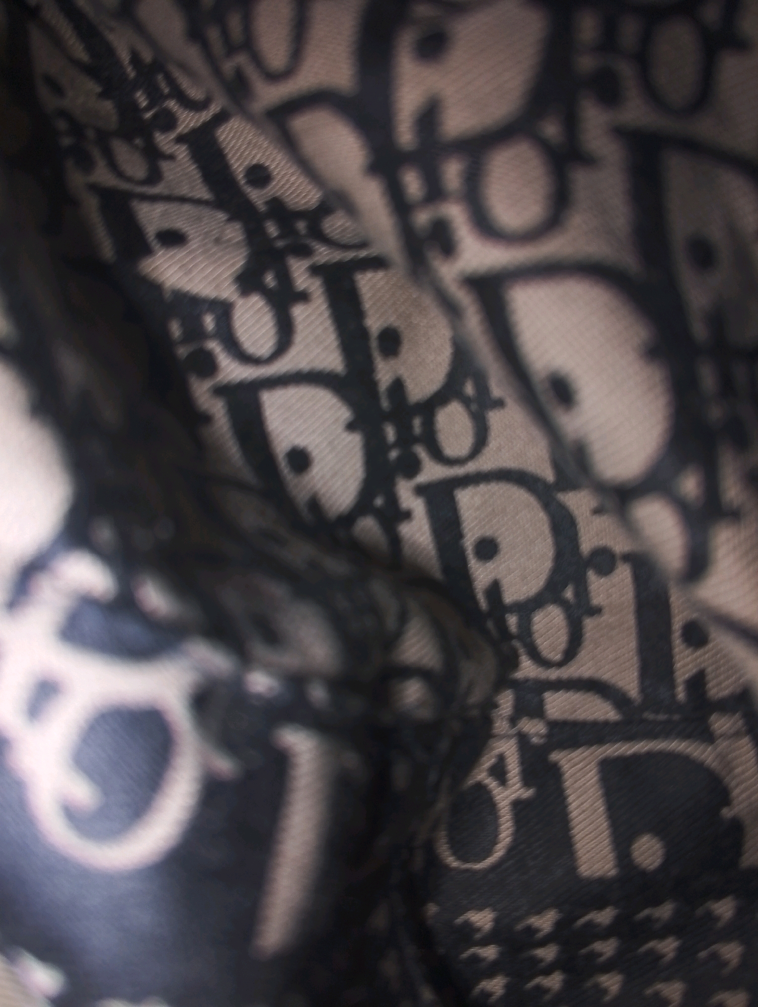 The image shows a close-up, blurry view of a fabric with a repeating pattern of black "Dior" logos on a beige background. The fabric is creased and folded, obscuring much of the pattern. There is no indication of location within the city of Sumberwono, Indonesia, or any people, actions, or interactions. The overall impression is one of abstract texture and pattern.