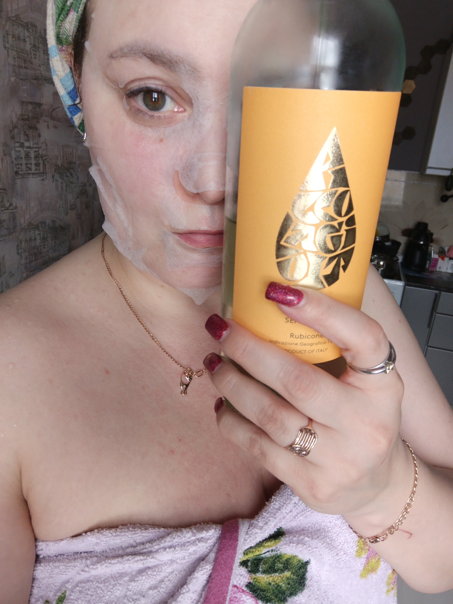 A woman wearing a sheet mask is holding a bottle with a yellow label and a golden teardrop-shaped design in front of her face. She is wearing a patterned headscarf, a gold necklace, and several rings and a bracelet. The lower half of her body is wrapped in a pink and white patterned towel. The label reads "RUBICONE" and "PRODUCT OF ITALY." The location is not visible and cannot be determined to be Марковский, Russia.