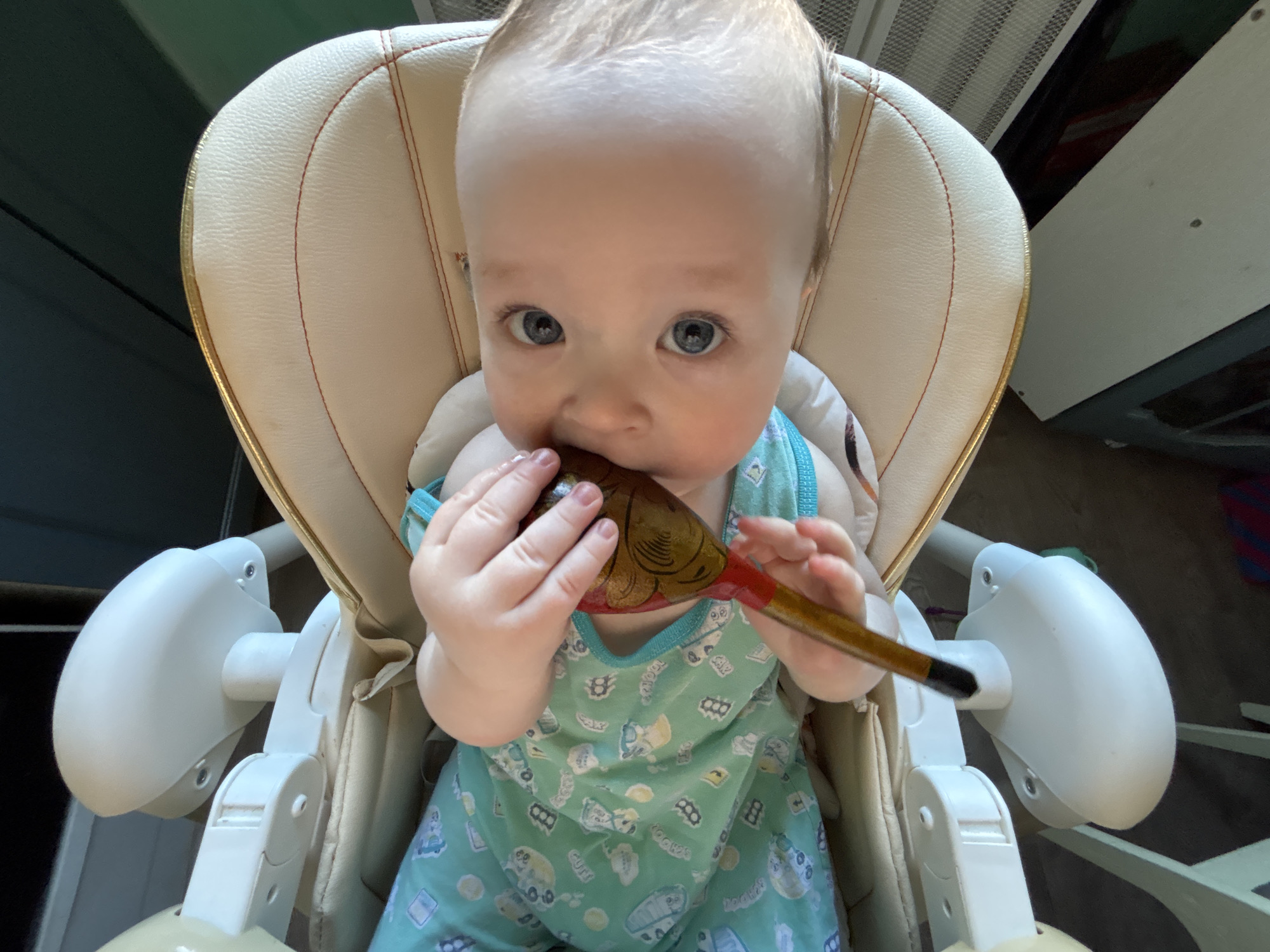 A baby is sitting in a high chair and chewing on a wooden spoon. The baby has light hair and blue eyes, and is wearing a light blue outfit with a cartoon pattern. The high chair has a white plastic frame and a beige padded seat and back. The background is blurry, but appears to be a room in a house. It is difficult to confirm that this image was taken in Марковский, Russia based on the visible elements.