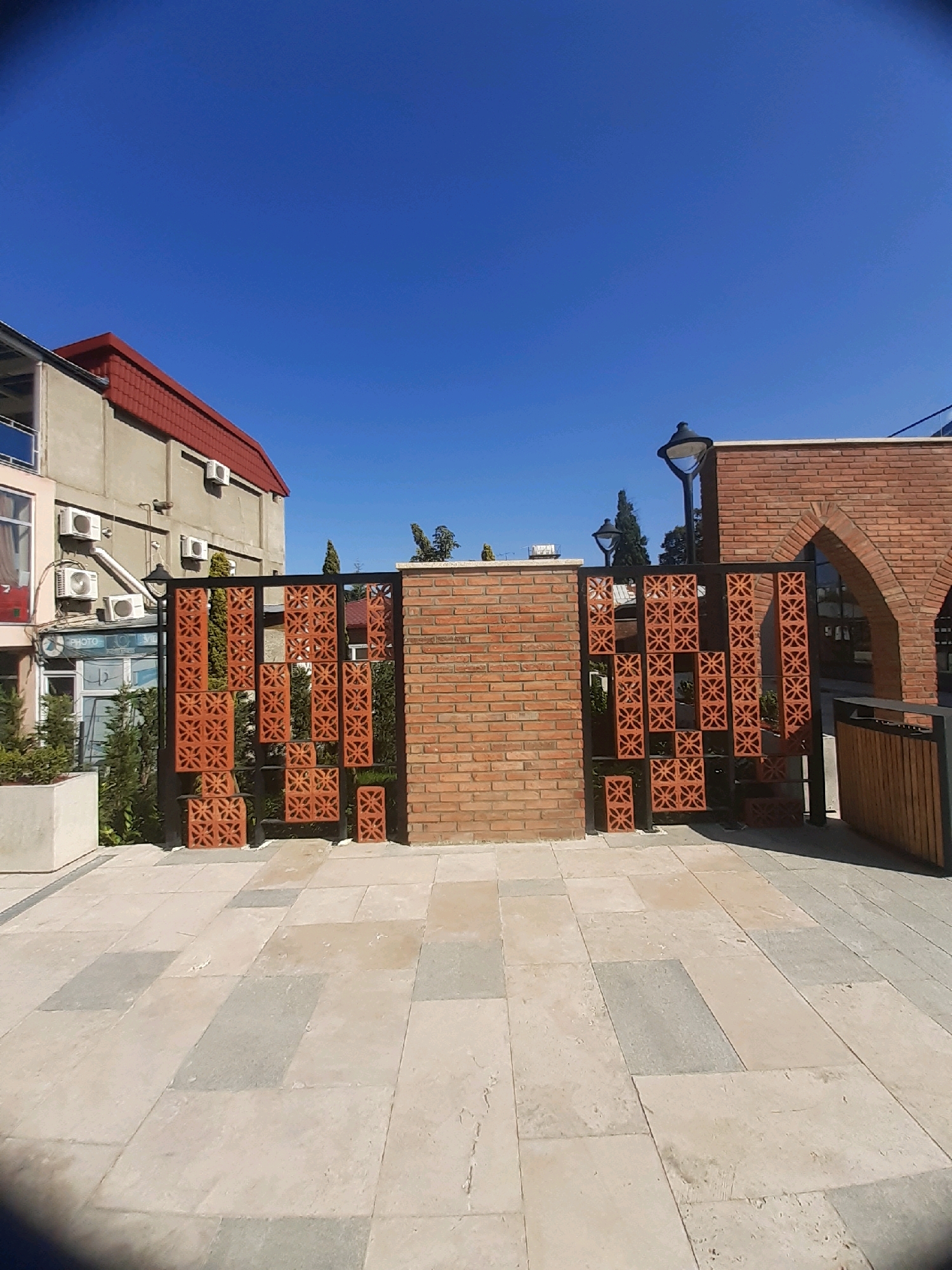 An outdoor daytime scene in Telavi, Georgia features a paved area with large, light-colored rectangular stones. A decorative brick wall with an arched opening and a wooden fence is visible on the right. In the center, a metal gate with a brick pattern is positioned in front of a solid brick wall. To the left of the gate, a building with a red-tiled roof and a concrete facade stands, adorned with several air conditioning units. The sky is a clear, bright blue.